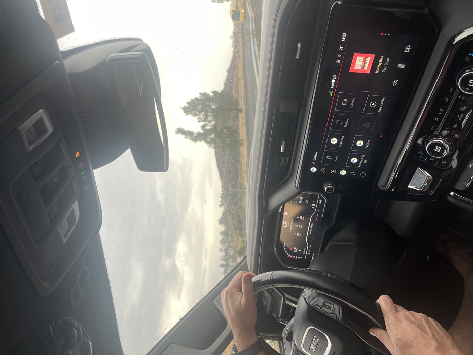 The image presents an interior view of a modern GMC vehicle, looking forward through the windshield at a scene in Santa Ana Acozautla, Mexico. The driver's hands, appearing masculine, are positioned on the steering wheel; a dark watch is visible on the left wrist. The digital instrument cluster displays a speed of "102" (likely km/h), a compass heading of "NE", and the time "15:32". To the right, a large digital infotainment screen shows multiple application icons, including "Google Maps" and "Apple CarPlay". Currently, the song "The Way Back" by Zach Bryan is playing, and the screen indicates the time as "14:32". The overhead console features active green and orange indicator lights (likely OnStar and passenger airbag status). The windshield has minor spots. Outside the vehicle, a road with a yellow guardrail is visible on the right. The landscape consists of low, rolling hills or mounds covered with sparse vegetation and scattered trees. The sky is uniformly overcast. In the distance, on the right side of the road, a portion of a yellow vehicle, possibly a bus, is discernible.