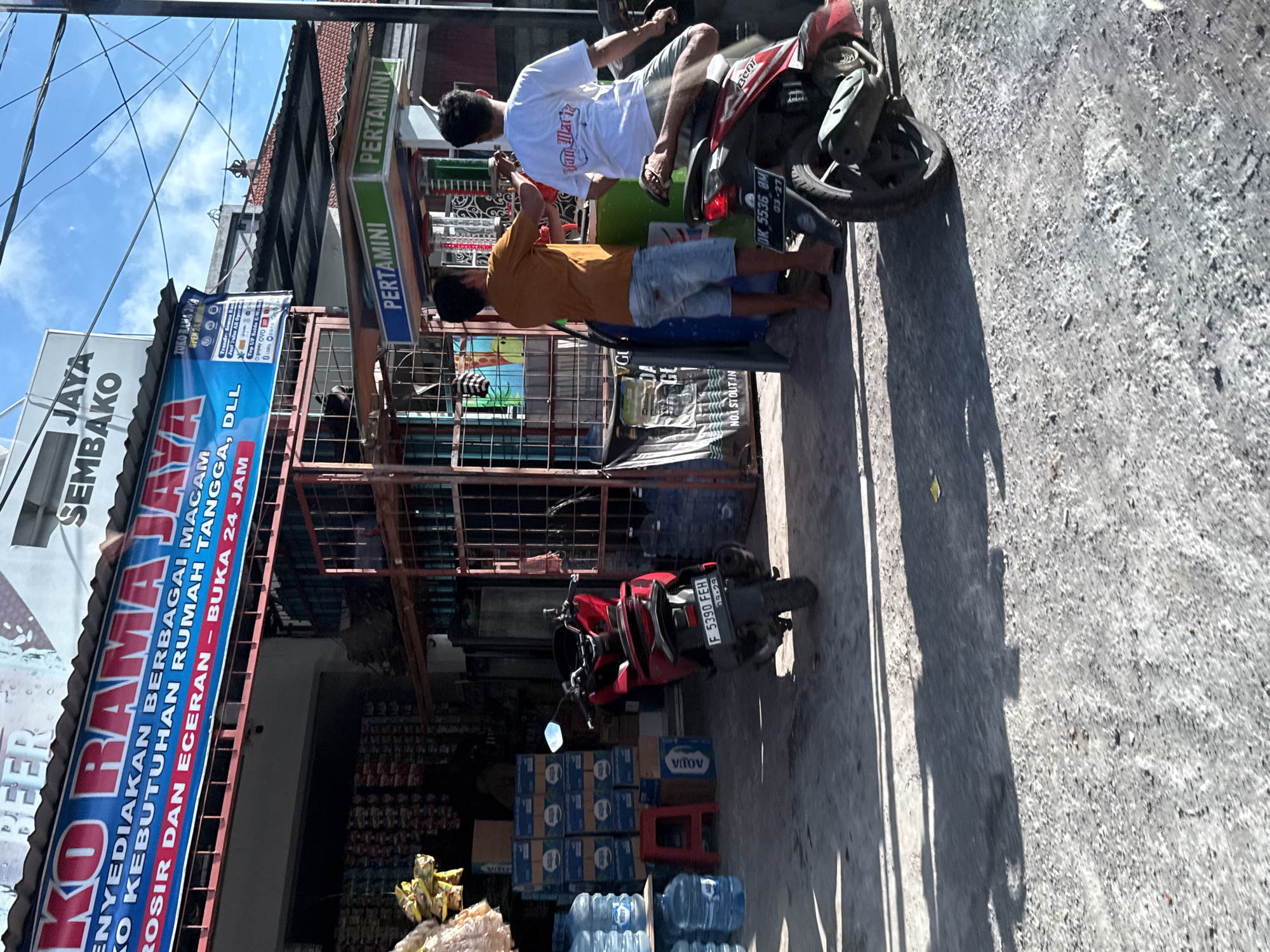 This image captures an outdoor scene in Kuta Selatan, Indonesia, depicting a "PERTAMINI" roadside fuel station operating alongside a convenience store under bright midday sun. Two men are featured with two motorcycles. One man, wearing a white t-shirt and grey shorts, sits on a red and black Honda Beat scooter (license plate DK 5536 BM, expiry 03-27). The other man, in a yellow/orange t-shirt and light blue shorts, appears to be operating one of the "PERTAMINI" fuel dispensers, possibly filling a container or the tank of a red scooter (license plate F 5390 FEH, expiry 09-29) parked closer to the store.
The adjacent shop is identified by banners as "JAYA SEMBAKO" and "KORAMADA". Visible text on the banners indicates it "MENYEDIAKAN BERBAGAI MACAM KEBUTUHAN RUMAH TANGGA, DLL" (provides various household needs, etc.) and offers "GROSIR DAN ECERAN - BUKA 24 JAM" (wholesale and retail - open 24 hours). Another banner for "TOKO RAMA JAYA" also states "OPEN 24 JAM" and lists services such as "Transkasi Semua Bank" (all bank transactions), "Jual Pulsa All Operator" (sell all operator credit), and "Top UP Saldo E-Wallet" including "Gopay OVO DANA QRIS ShopeePay". Large bottles of water, some stacked in crates, are visible outside the shop, along with other general merchandise. The clear blue sky and distinct shadows suggest a sunny day.