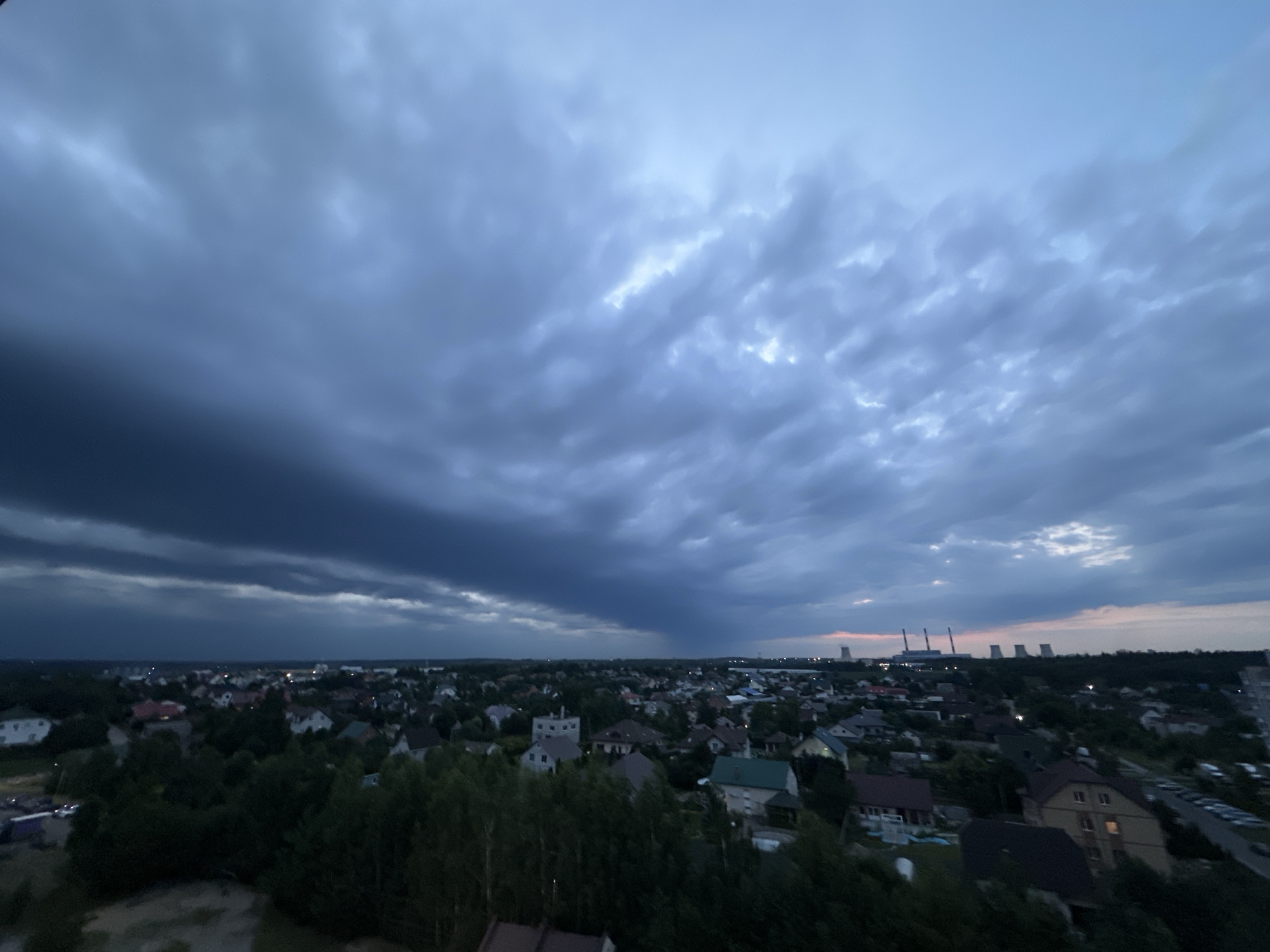 This is a wide-angle, high-angle shot of a residential area in Шчомысліцкі сельскі Савет, Belarus, at dusk. The sky is filled with heavy, dark, and dramatic clouds, suggesting an approaching storm or recent rainfall. The clouds are tinged with blues and grays, with some lighter patches indicating diffused light. Below the sky, a sprawling suburban landscape unfolds, characterized by numerous houses with pitched roofs, surrounded by dense green trees. The houses appear to be of varying sizes and architectural styles, with some having visible windows and chimneys. In the distance, a line of industrial structures with tall chimneys, likely a power plant, is silhouetted against the slightly brighter horizon. The overall impression is one of a vast, somewhat somber, and natural setting at the edge of a populated area. There are no people or vehicles clearly visible in the scene.