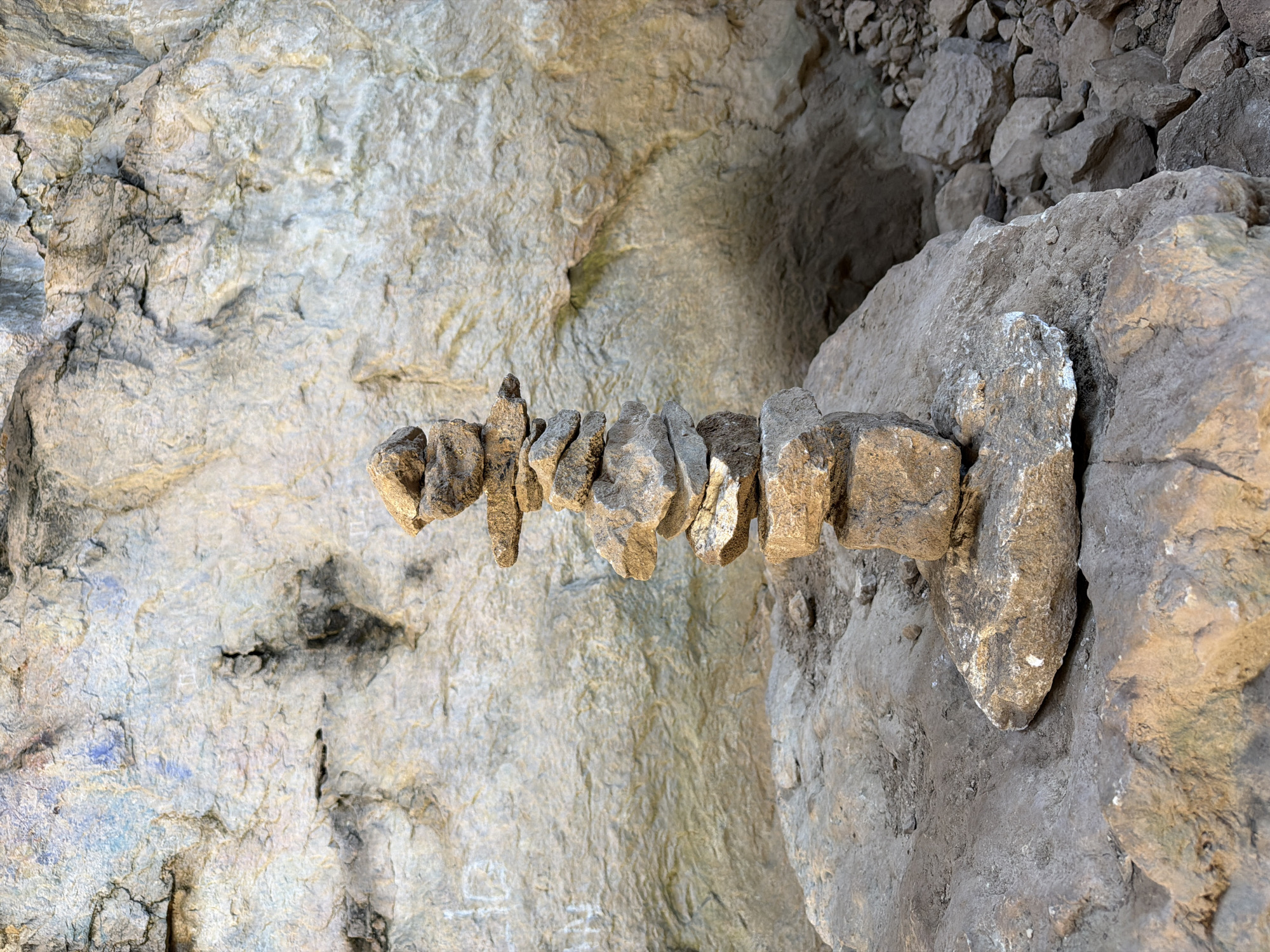 A geological formation resembling a rock bridge or shelf protrudes horizontally from the face of a rock wall. The formation appears segmented or constructed from stacked stones, extending from a larger, more solid rock mass. The rock face is textured and light in color. Debris and loose rocks are visible in the background. The location is in the city of Karlukovo, Bulgaria.