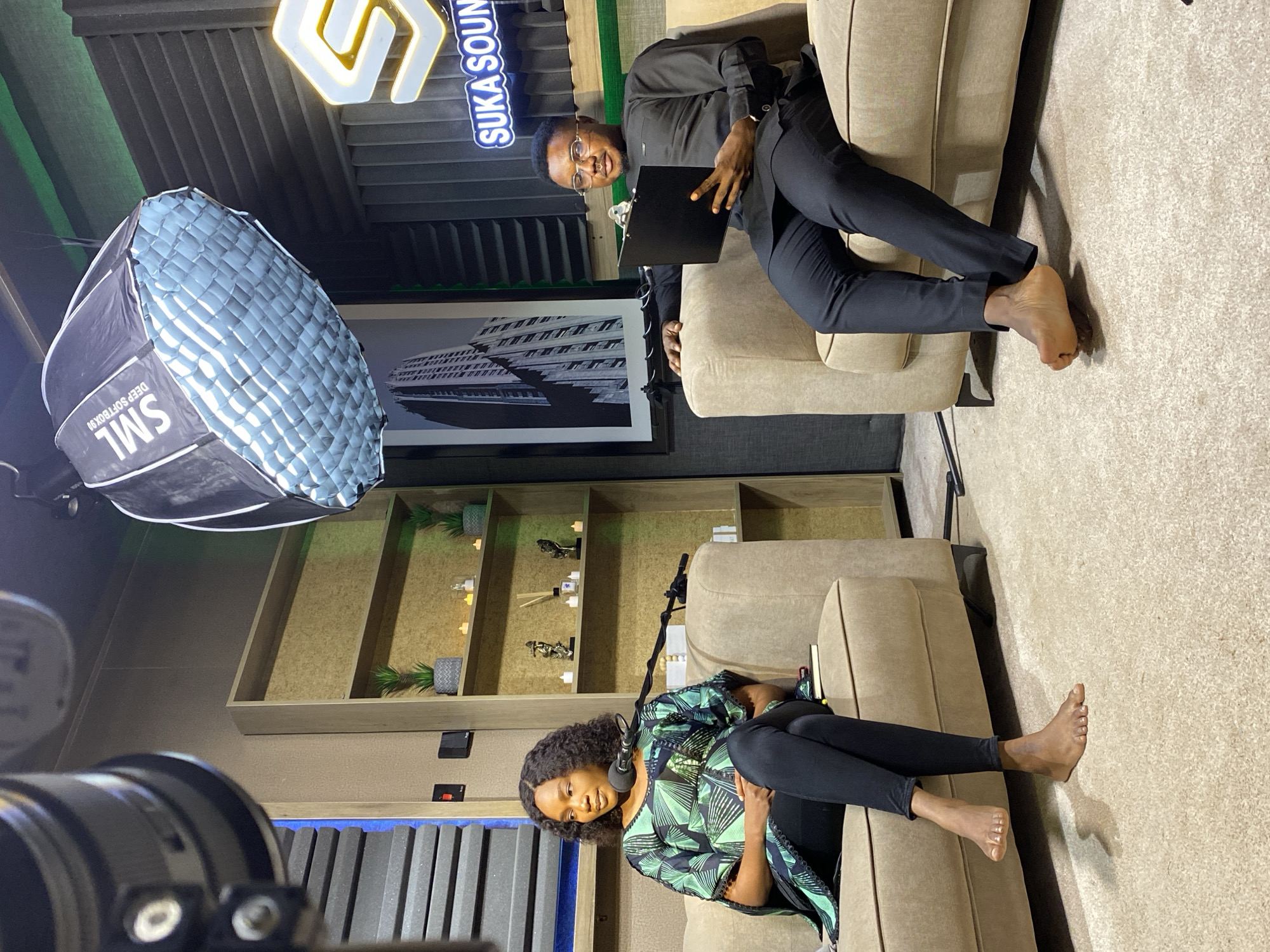 Two individuals are seated on a tan sofa in a recording studio. The man, on the left, is wearing a black collared shirt and black pants. He has glasses and is holding a black clipboard. He is barefoot. The woman, on the right, is wearing a green and black patterned top and black pants. She is also barefoot. A microphone is positioned in front of her.
Behind the subjects, shelves are visible, adorned with decorative items and small potted plants. A large, octagonal softbox light is positioned to the left of the frame. In the background, a neon sign with the text "SUKA SOUND" is illuminated. A framed black and white photograph of a tall building hangs on the wall. The studio has acoustic paneling on the walls and a carpeted floor. The lighting suggests it is indoors, likely evening or night. The overall atmosphere appears to be set for a recording session or interview.