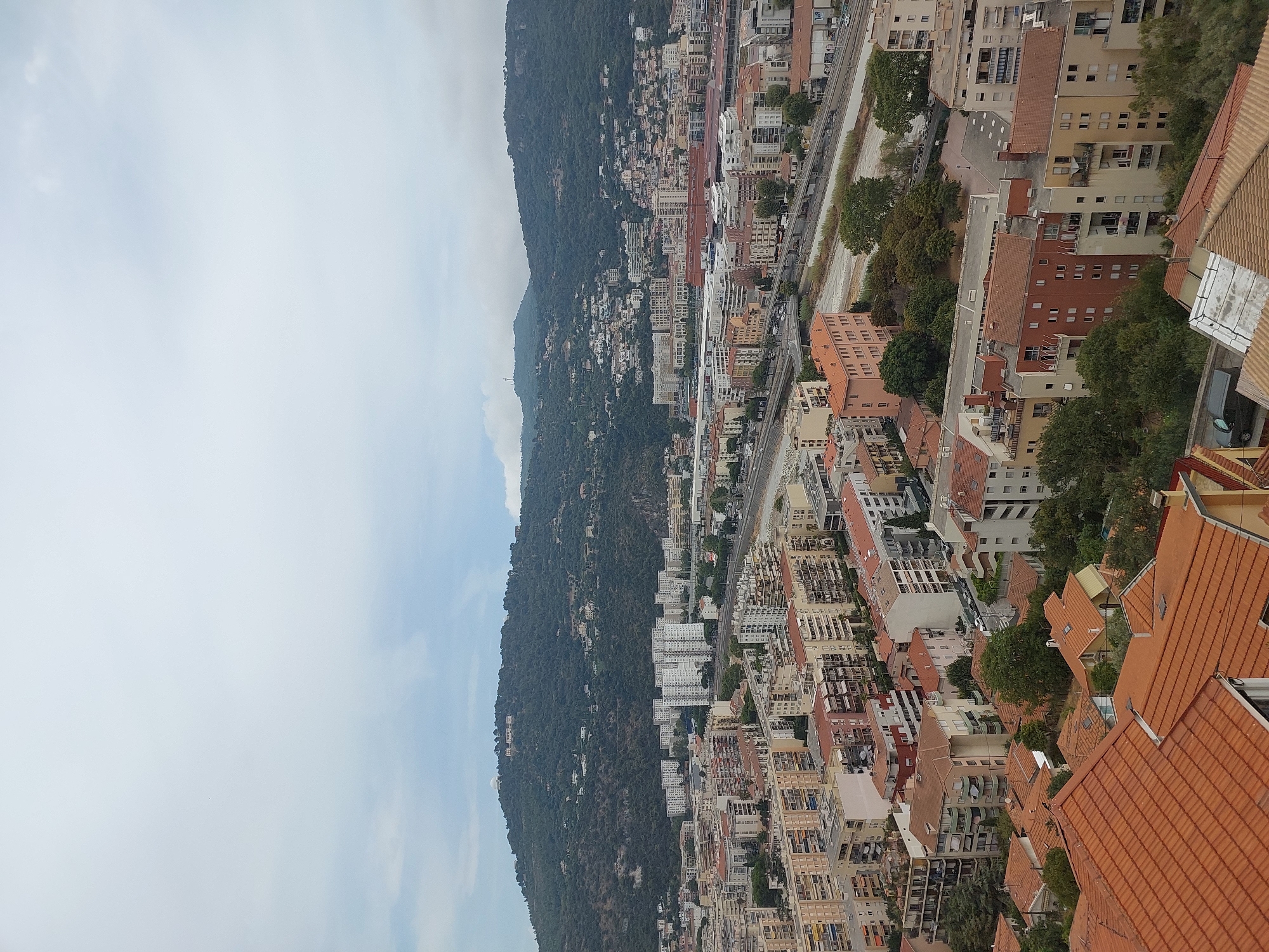 This is an aerial, medium-angle, eye-level shot of a densely populated urban environment in Nice, France. The foreground features a cluster of buildings with terracotta tile roofs, typical of Mediterranean architecture. These buildings range from single-family homes to multi-story apartment complexes, many with balconies.
Beyond the foreground, the cityscape expands, revealing a mix of residential and commercial structures. Numerous apartment buildings, primarily white or pastel-colored, dominate the middle ground. A road or highway runs horizontally through the scene, with cars visible on it, suggesting active transportation. Adjacent to the road, a dry riverbed or canal is visible.
In the background, a large, tree-covered hill rises, dotted with smaller houses and structures, indicating development on the hillside. The sky is overcast with clouds, casting a diffused light over the entire scene. The overall impression is of a sprawling coastal city nestled against a natural, mountainous backdrop, under a cloudy sky. No people or animals are clearly discernible in the image.