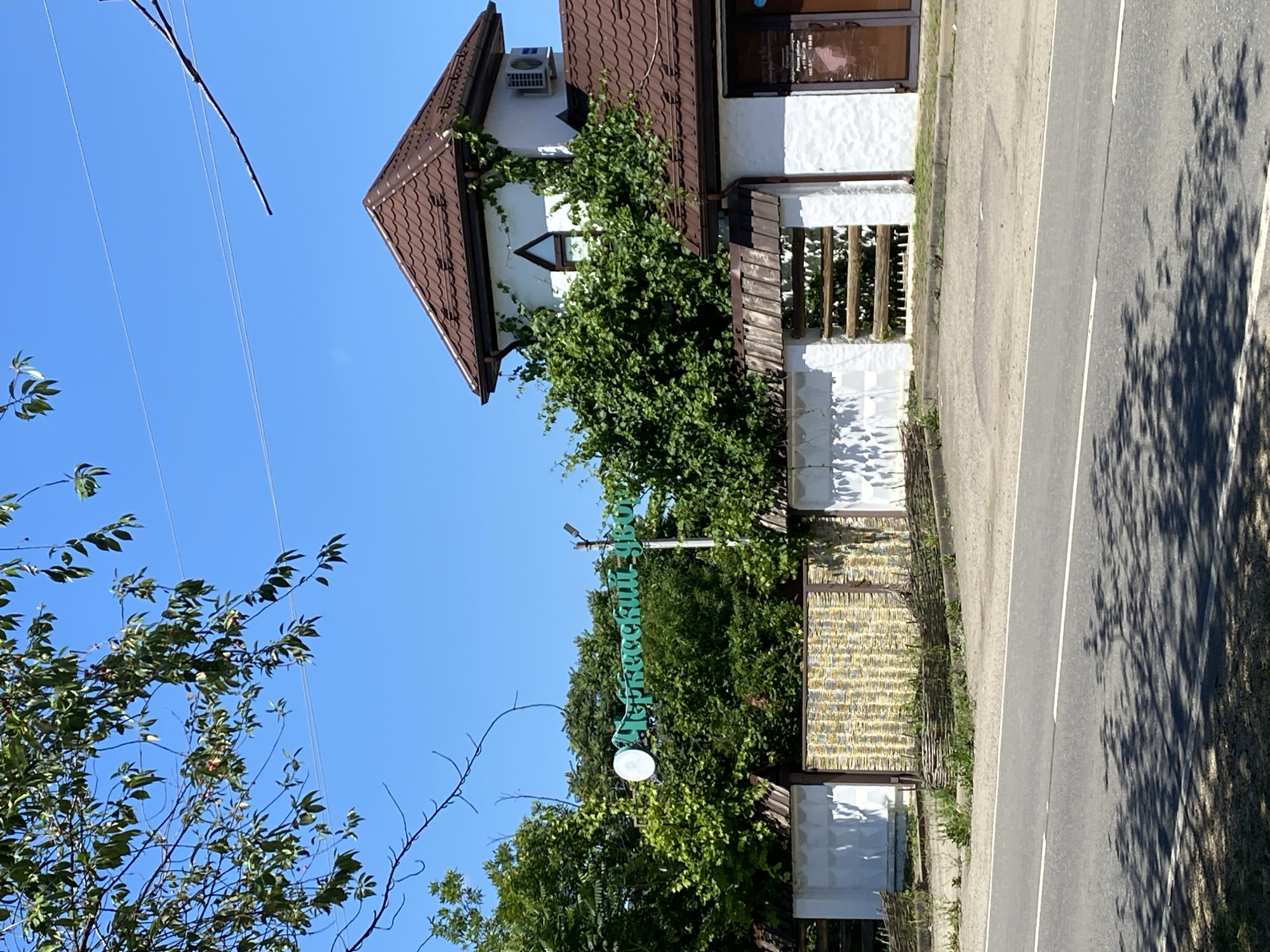 The image depicts a sunny day scene in Zapadny, Russia. A white stucco building with a brown tiled roof is visible, partially obscured by lush green foliage and vines. A tower-like structure attached to the building features a small triangular window and an air conditioning unit. In front of the building, a white fence with a decorative woven section runs along a paved road. A green sign with Cyrillic lettering is mounted on a pole behind the fence, amidst the trees. The road in the foreground is asphalt, marked with a white line. The overall environment suggests a commercial or residential property surrounded by vegetation under a clear blue sky.