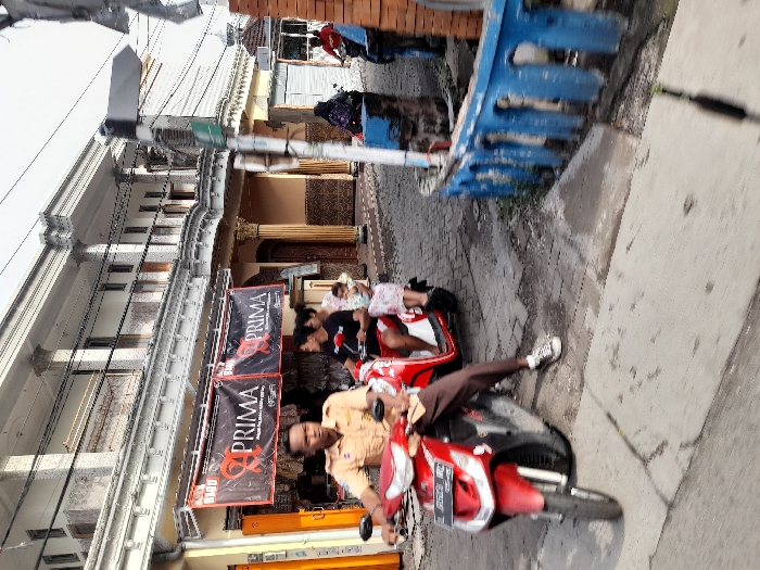 On a street, a man wearing a tan shirt rides a red motorcycle. Behind him on another red motorcycle are a man and a child. Banners hang on a building. There is another motorcycle in the background with what seems to be a child on it. The image is tilted.