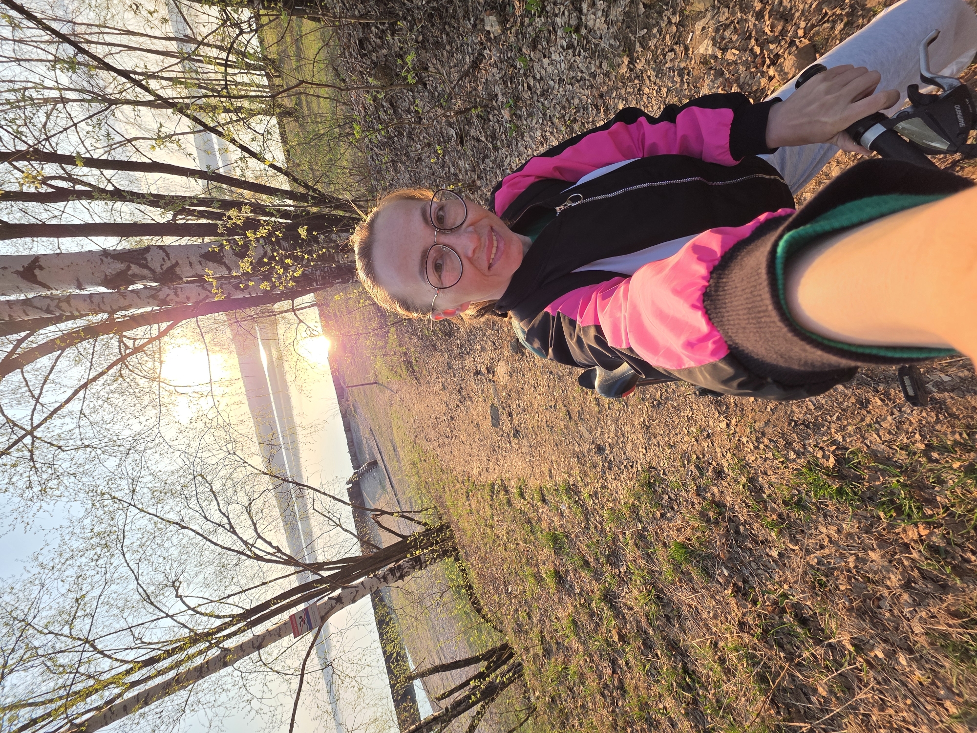 A woman is pictured in an outdoor setting in what appears to be the city of Chaikovsky, Russia, based on visual information. The woman, wearing glasses and a black jacket with pink accents, is positioned at an angle, likely taking a selfie. She is holding the handlebars of a bicycle. The background includes trees, a body of water, and the sun setting, indicating the location is near a river or lake. The ground is covered in dry leaves and some patches of grass. The overall impression is a casual outdoor moment captured on camera.