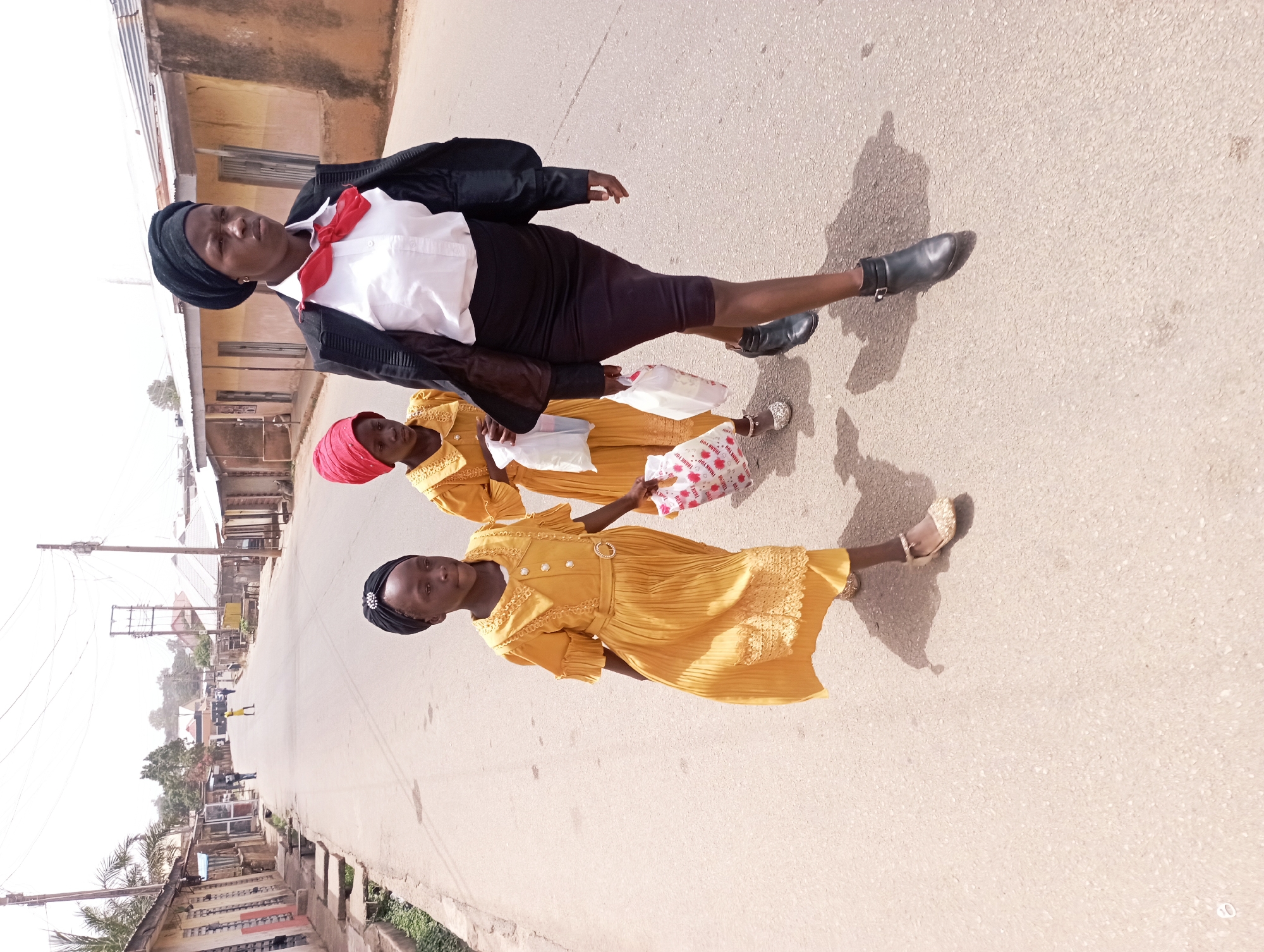 The photograph shows three individuals walking on a paved street in Jos, Nigeria. The person in the front is wearing a black blazer, white collared shirt, red bow tie, black skirt, black head covering, and black shoes. The two individuals behind are wearing yellow dresses with red head coverings and carrying what appears to be paper shopping bags. The buildings on the sides of the street are a mix of residential and commercial structures. Utility poles are visible along the street. The photograph appears to be taken during the daytime.