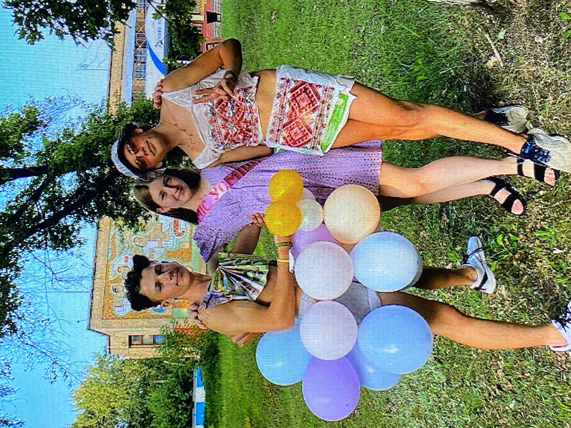 Three individuals are posing for a photo outdoors, in front of a building with a mural. The person on the left, a male, is wearing a sleeveless top adorned with a colorful fabric scrap and grey shorts. He is holding a bouquet of pastel-colored balloons. The person in the middle, a female, is wearing a short lavender dress with a pink sash and is also holding yellow balloons. The person on the right, a male, is wearing a white top with red embroidery, a short skirt with similar embroidery, a bucket hat, and sunglasses. He is making a peace sign with his hand. The background shows a grassy area with trees and a building featuring a large mural. The overall setting appears to be a park or school grounds on a sunny day. There is no information in the image to suggest this location is in Poznan, Poland.