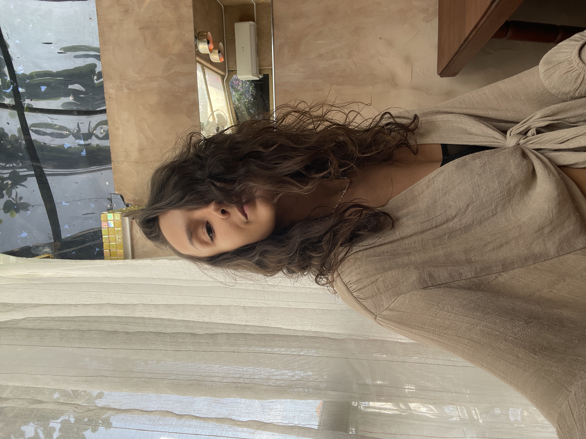 A medium shot captures a woman with long, dark, wavy hair looking directly at the camera. She is wearing a loose-fitting, light beige or tan top, possibly made of linen or a similar fabric, which is tied in a knot at the front, revealing a glimpse of a dark undergarment. A delicate, light-colored necklace is visible around her neck.
The setting appears to be an indoor space, likely a room with a window and a mirror. To her left, behind her, is a window covered by sheer, light-colored curtains that diffuse the natural daylight. Through the window, faint green foliage is visible. To her right, a warm-toned, possibly tiled or textured wall extends, with a mirror reflecting elements of the room. In the mirror's reflection, a small white air conditioning unit is mounted, and some cylindrical, orange-toned decorative or functional items are seen on the wall. Another reflection in the mirror shows what appears to be more greenery or an outdoor scene. The overall lighting is soft and natural, suggesting it is daytime. The woman's expression is neutral and calm. There is no visible text in the image. The scene conveys a relaxed atmosphere in a location like Mũi Né, Vietnam.