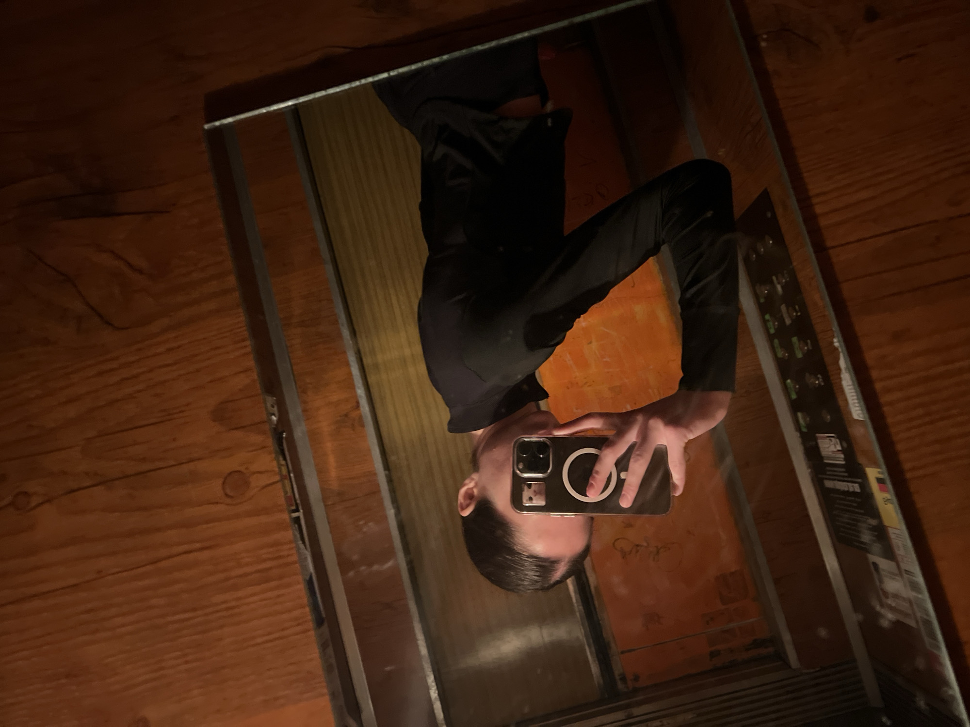 A reflection of a person holding a phone in an elevator is captured in the image. The person is wearing dark clothing and appears to be taking a selfie. The elevator interior is visible, with wooden paneling on the floor, a control panel with buttons, and vertical metal frames. The image appears to be taken at an angle, and the reflection is slightly distorted. It is not possible to identify the exact location or city from the image alone without additional information.