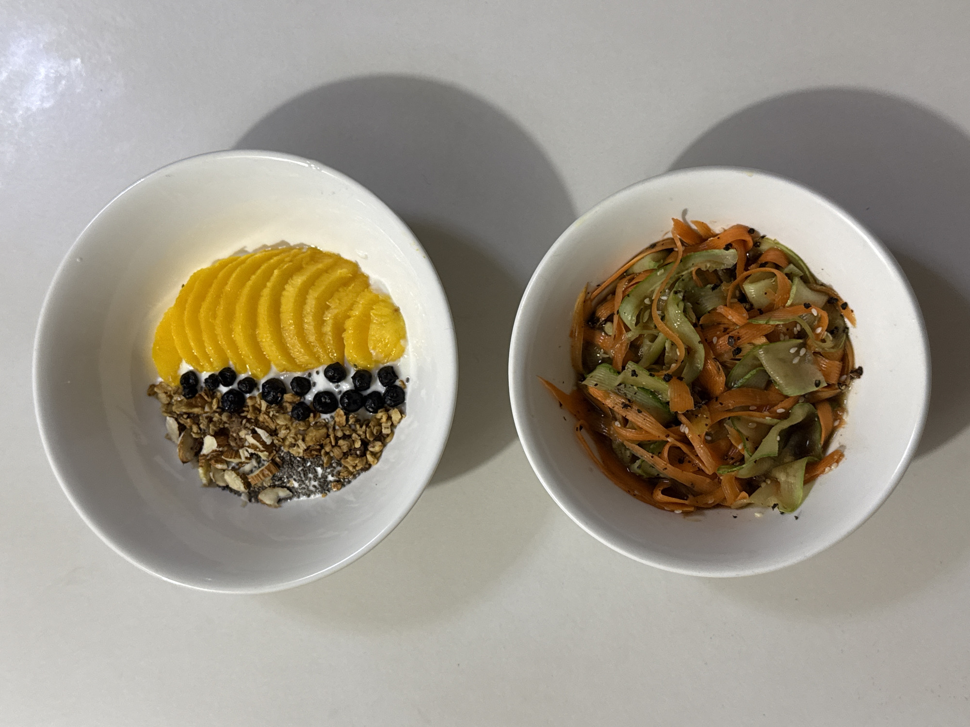 The scene is situated in Davao City, Philippines.
Two white ceramic bowls of food are presented on a smooth, light gray surface, illuminated from the upper left, casting distinct shadows to the lower right of each bowl.
The upper bowl contains a layered food item. A white base, likely yogurt, is visible beneath toppings arranged in sections. On the left, a mixture of brown granola, chopped almonds, and black chia seeds rests. Adjacent to this, a vertical line of small, dark blueberries is visible. To the right, a fan-like arrangement of bright yellow mango slices completes the toppings.
The lower bowl holds a mixed vegetable salad or slaw. It consists of thin, wavy ribbons of vibrant orange carrots and pale green cucumbers. The vegetables are coated in a subtle, glistening dressing and garnished with black specks, possibly ground pepper, and scattered white sesame seeds.
No individuals are visible in the image.