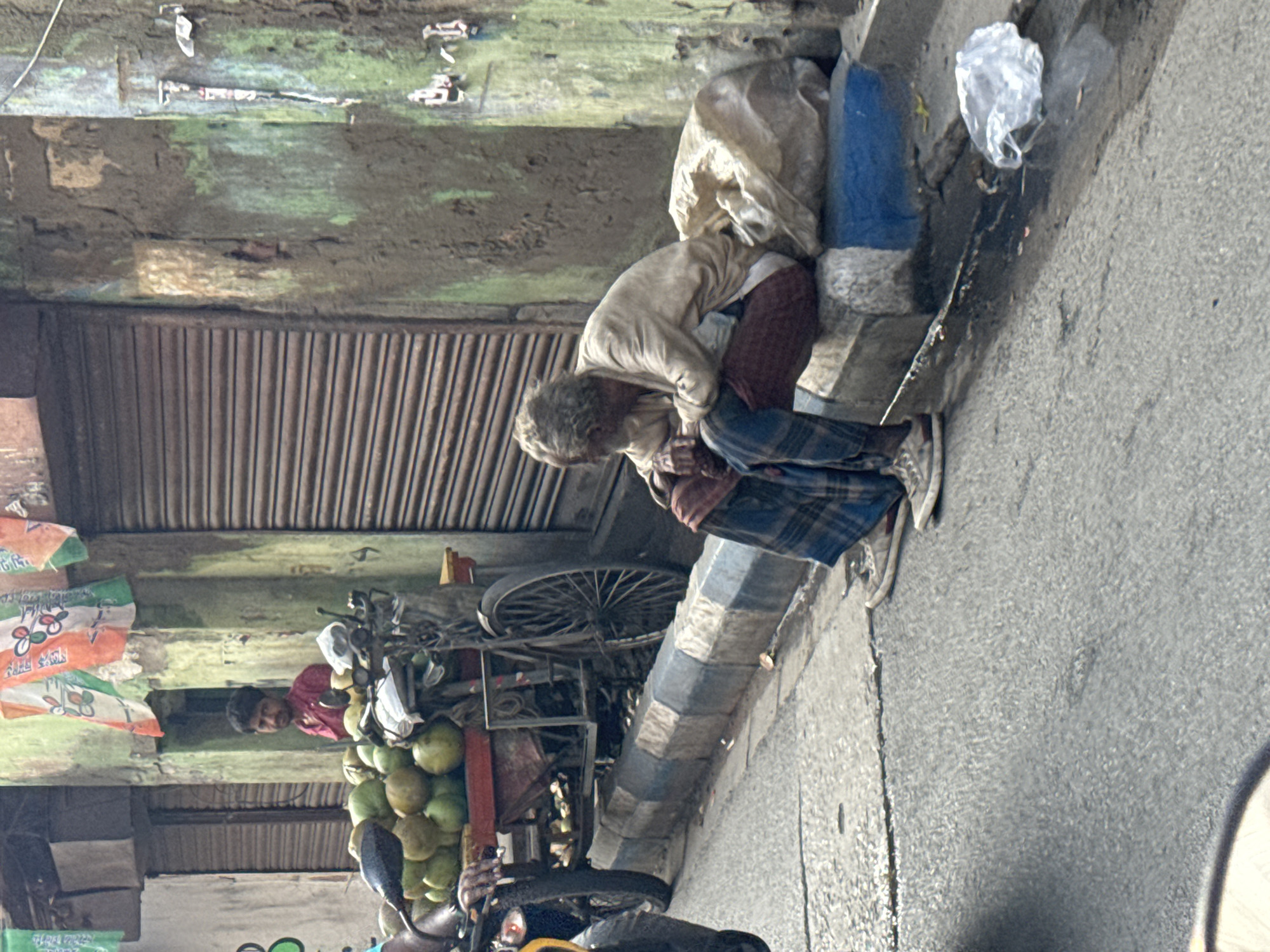 An elderly man, gray-haired and dressed in worn clothing, is squatting on the curb of a street in Kolkata, India. He wears a light-colored shirt and plaid pants. Behind him, a corrugated metal shutter is partly closed, and the adjacent wall shows signs of decay with greenish stains. A bicycle cart loaded with coconuts stands in front of the shutter, and a man in a red shirt stands behind it. Debris, including plastic bags, are visible on the street. The sidewalk is gray and marked with dark, streaky stains.