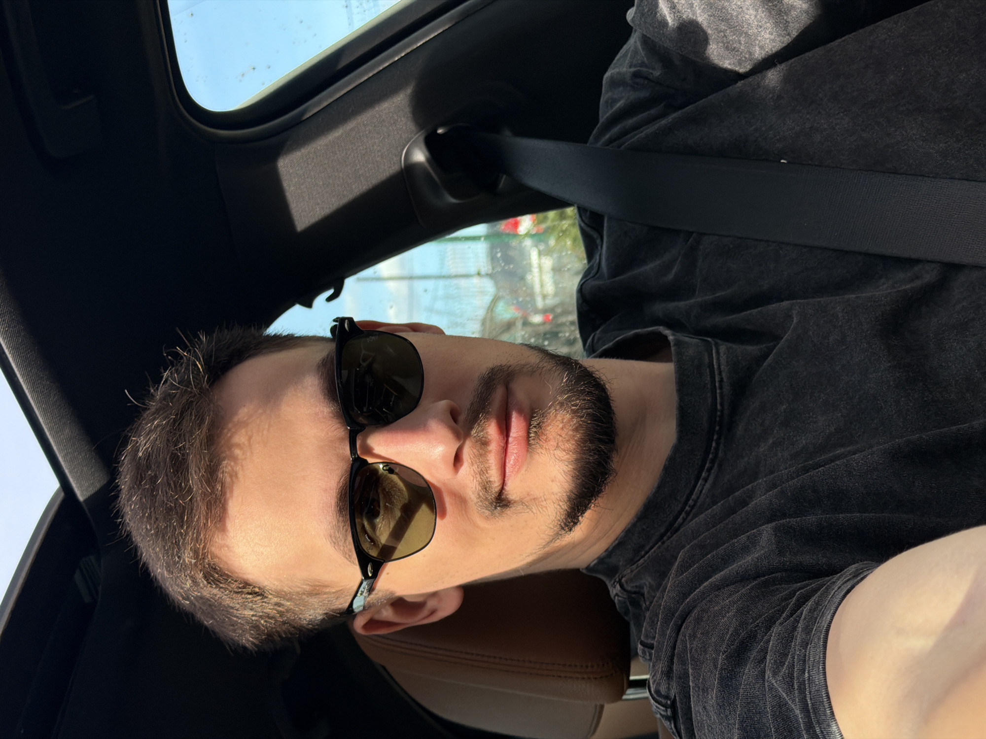 A man is in a vehicle, seemingly a car, as evidenced by the car's interior visible including the sunroof, safety belt and interior roof. The man has light colored skin, short brown hair, a beard, and is wearing sunglasses. He is wearing a gray shirt. He is likely driving or in the passenger seat of a vehicle. It is daytime, based on the amount of light. There is no evidence in the photo indicating location in Krasnoyarsk, Russia.