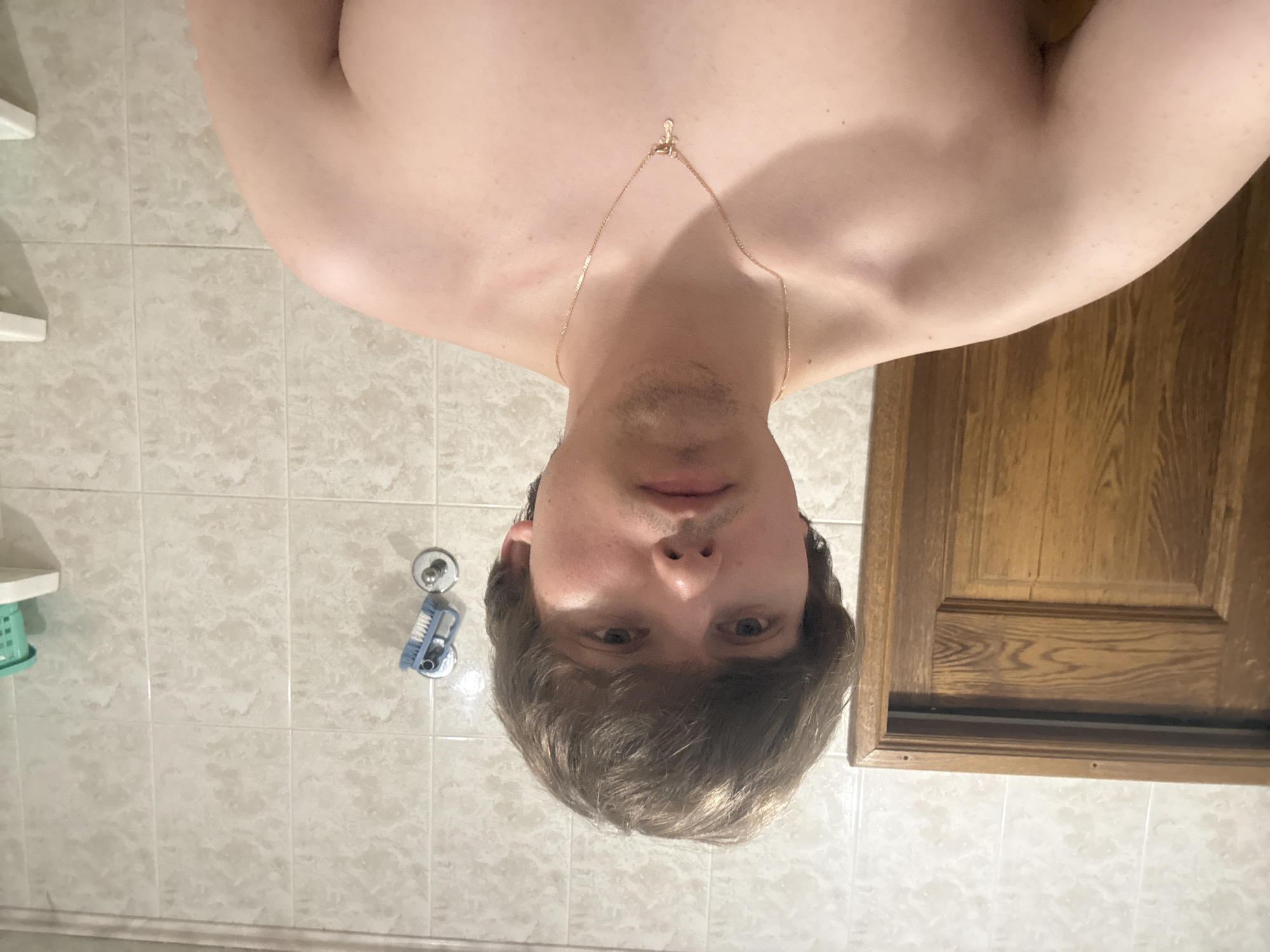 The image shows a chest-up, front-facing portrait of a young, shirtless Caucasian male with light skin and light brown hair. He is wearing a thin, gold chain with a small, cross-shaped pendant. His facial expression is neutral, and he has a light stubble on his chin and upper lip. The background consists of tiled walls in a bathroom, a wooden door on the left, and a shelf with a green plastic container on the right. A blue cleaning brush is attached to the wall with a suction cup to the right of the man's head. The lighting appears to be artificial and somewhat harsh, casting subtle shadows. The scene is indoors, likely in a residential bathroom. The location is identified as Dnipro, Ukraine.