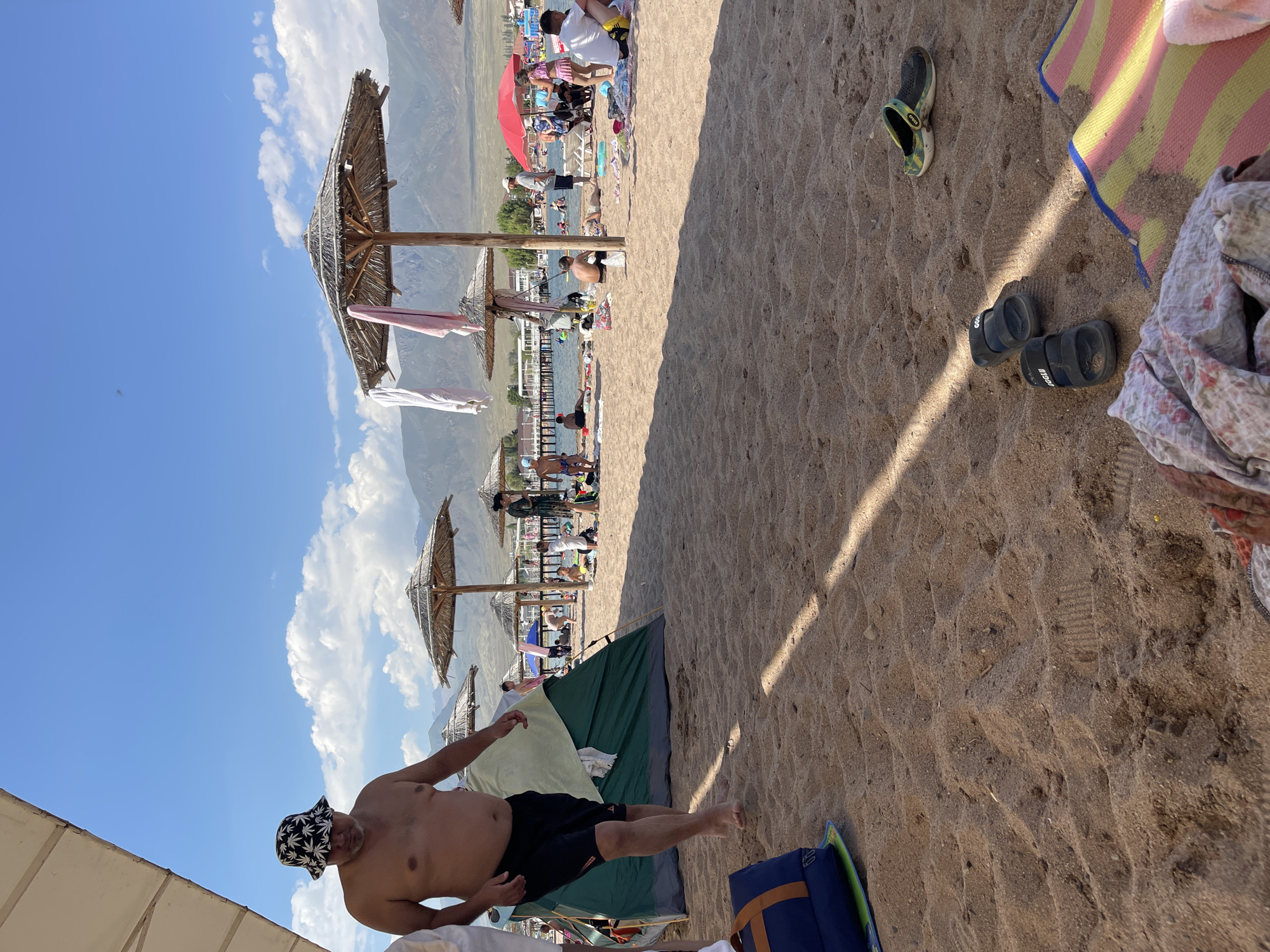 A busy beach scene unfolds under a bright, partly cloudy sky with distant mountains. In the foreground, a man wearing a bucket hat and black swim trunks walks on the sand towards the left. A green tent is set up behind him, with a blue and orange bag on the sand to his left. The beach is populated with numerous people sunbathing, sitting on towels, and walking around. Several thatched umbrellas provide shade for beachgoers. In the background, there is a body of water, possibly a lake, with a pier extending into it. The overall environment suggests a popular tourist destination on a warm day in Choktal, Kyrgyzstan.
