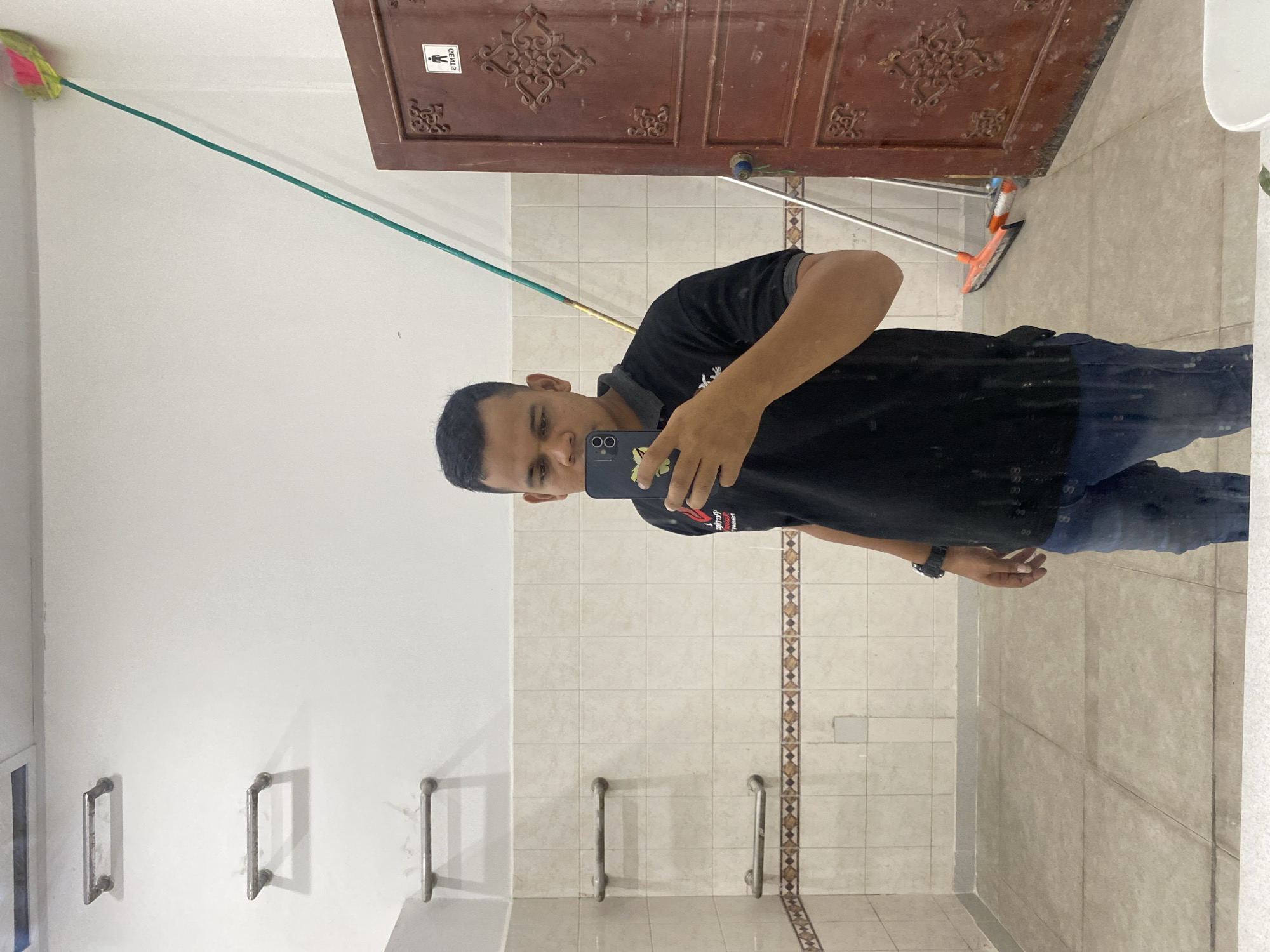 A man takes a mirror selfie in a tiled room, possibly a public restroom or changing facility, in Banda Aceh, Indonesia. He is wearing a black polo shirt and blue jeans. In the background, there are several metal grab bars mounted on the wall. A broom and a mop stand near a wooden door with ornate carvings and a "GENTS" sign. The room is brightly lit, and the walls and floor are tiled. The man is holding a smartphone and appears to be looking at its screen.