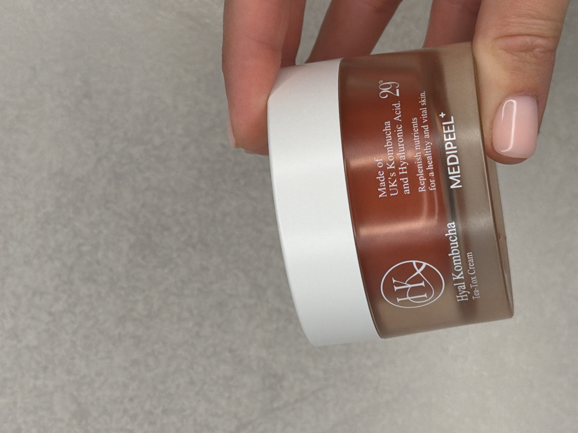A person's hand is prominently featured, holding a cosmetic product jar. The hand's fingers are wrapped around the container, showcasing neatly manicured nails with a glossy, light pink or nude polish.
The object is a jar of "MEDIPEEL+" branded cosmetic cream, specifically "Hyal Kombucha Tea-Tox Cream". The jar has a transparent, brownish-amber body and a solid white lid. Visible text on the label includes a logo with a stylized "K", "Hyal Kombucha", "Tea-Tox Cream", and details about its ingredients and purpose: "Made of UK's Kombucha and Hyaluronic Acid.", "29°", and "Replenish nutrients for a healthy and vital skin.". The brand name "MEDIPEEL+" is also clearly displayed.
The scene takes place indoors, with the jar held against a plain, light grey, subtly textured surface, likely a tabletop or counter. There are no other objects or distinct features in the background. The lighting is soft and even, providing no specific time of day cues. The provided location context for this media file is Sofiivska Borshchahivka, Ukraine.