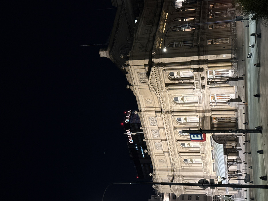 An ornately designed, illuminated building stands against a dark night sky. It features multiple rows of arched windows and decorative architectural details. A sign with the word "QUALIA" in red letters is visible above the building. A street with bollards lines the right side of the image, and streetlights are also present.