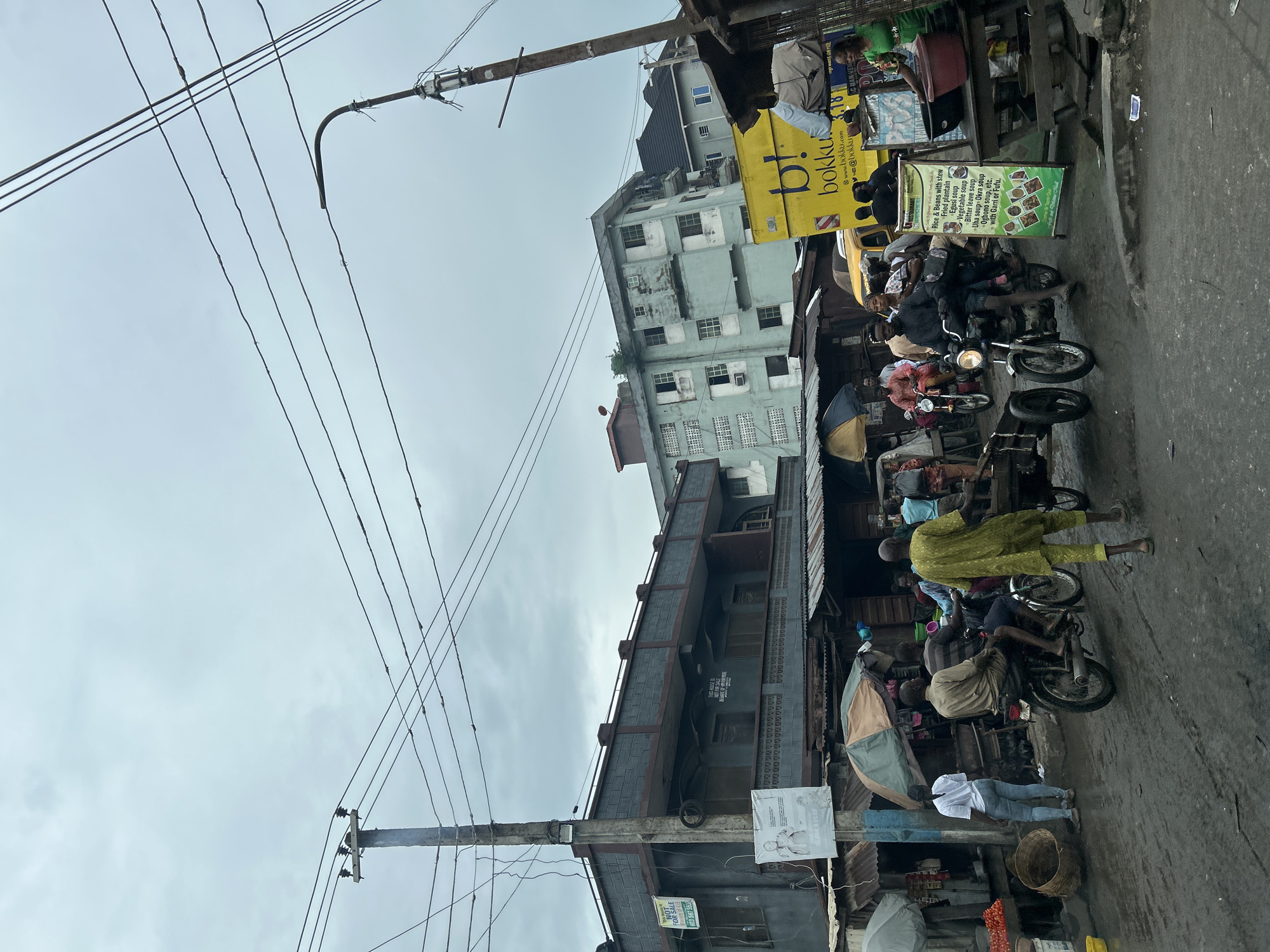 This is a street-level, medium-shot photograph taken in Lagos, Nigeria, likely during the day under an overcast sky. The scene depicts a bustling street with various people and vehicles, characteristic of a market or commercial area. In the foreground, a man in a bright green kaftan walks away from the camera, pushing a handcart. To his left, two men are riding on a motorcycle. Several other motorcycles and a yellow truck with "bokku.com" branding are visible in the middle ground.
The environment is a mixture of old and modern buildings. On the left, a dark grey, two-story building with a balcony and several windows stands prominently. A utility pole with numerous wires stretches across the frame in front of this building. Adjacent to it are makeshift stalls with corrugated metal roofs, selling goods. On the right side of the frame, a lighter blue, multi-story apartment building is visible. The street appears wet, suggesting recent rain, with visible tire tracks in the mud.
People are engaged in various activities: riding motorcycles, walking, and standing near stalls. The atmosphere appears dynamic and busy, with a mix of commercial and residential elements. The overall impression is of a vibrant urban environment in Lagos.