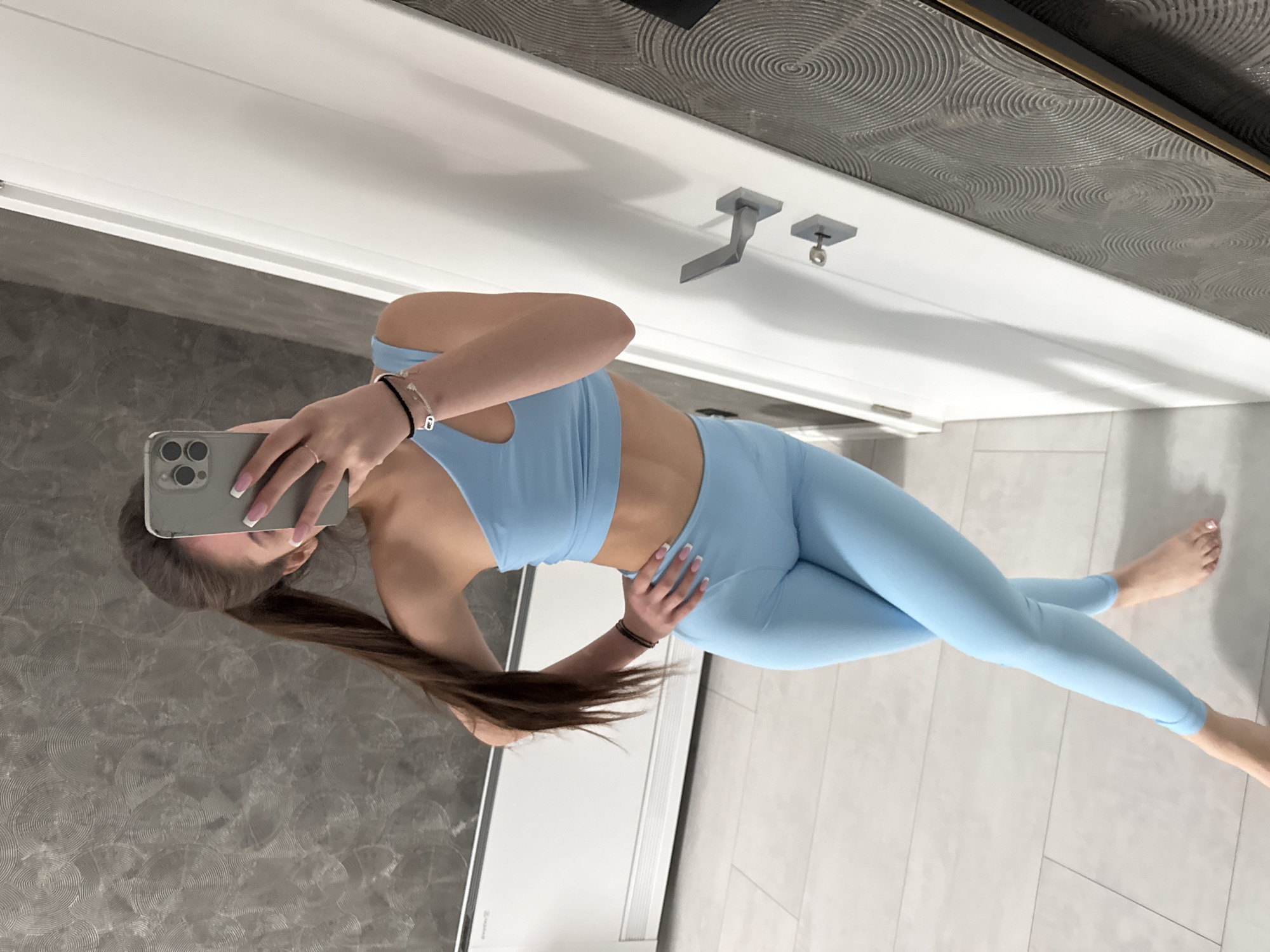 A person wearing a light blue athletic outfit takes a selfie in front of a mirror. The background is a room with white door and gray wall. The location is in Plovdiv, Bulgaria.
