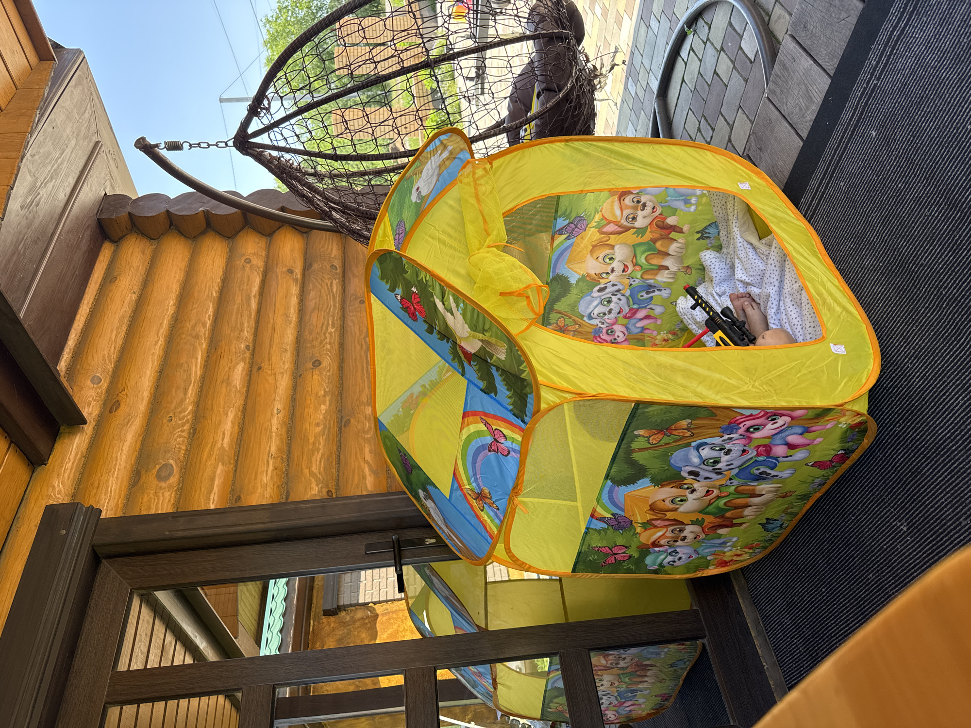 A child's feet are visible inside a yellow play tent. The tent is decorated with cartoon characters, possibly from the show Paw Patrol, and butterflies. Outside the tent, there is a dark-colored wicker hanging chair and a wooden-paneled building. The ground is paved with interlocking bricks and a dark, ribbed mat. The scene appears to be an outdoor porch or patio area. The image was taken in Minusinsk, Russia.