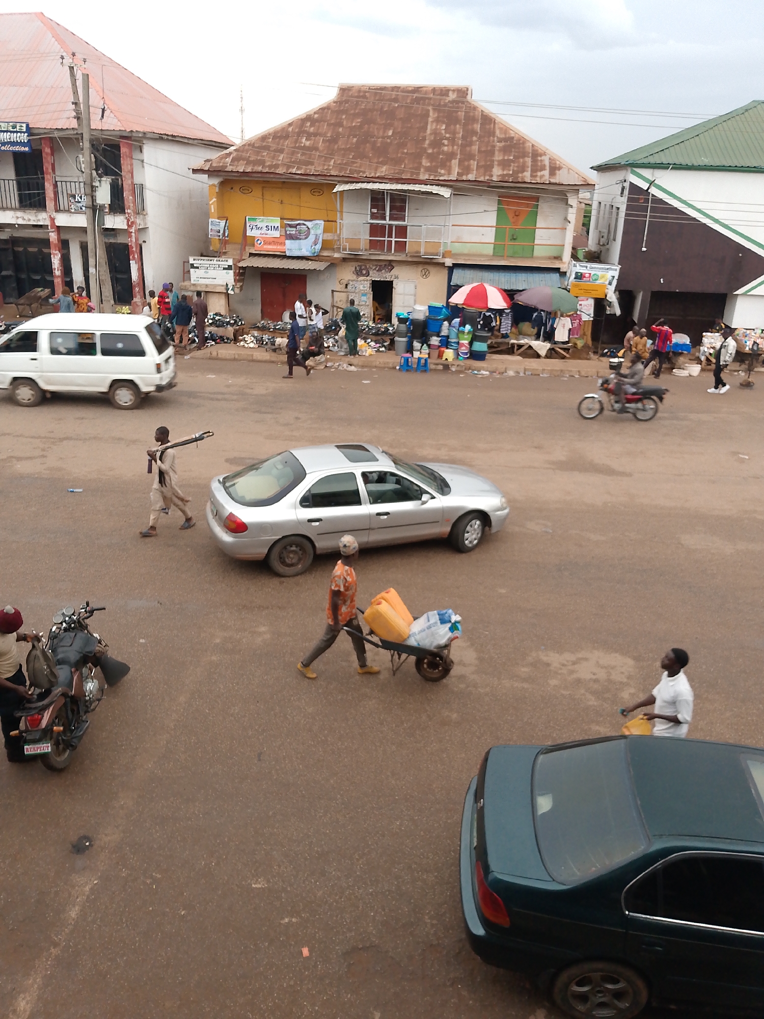 The image depicts a street scene in Bukuru, Nigeria. The road surface is reddish-brown, likely laterite soil. There are several buildings visible in the background, a mixture of one- and two-story structures, some with corrugated iron roofs. A small market area is present in the center, with people gathered around stalls and merchandise displayed. Vehicles present include a silver sedan, a dark green sedan, a white van, and motorcycles. People are walking across the road, some carrying items, one pushing a wheelbarrow with water containers. The sky is overcast.
