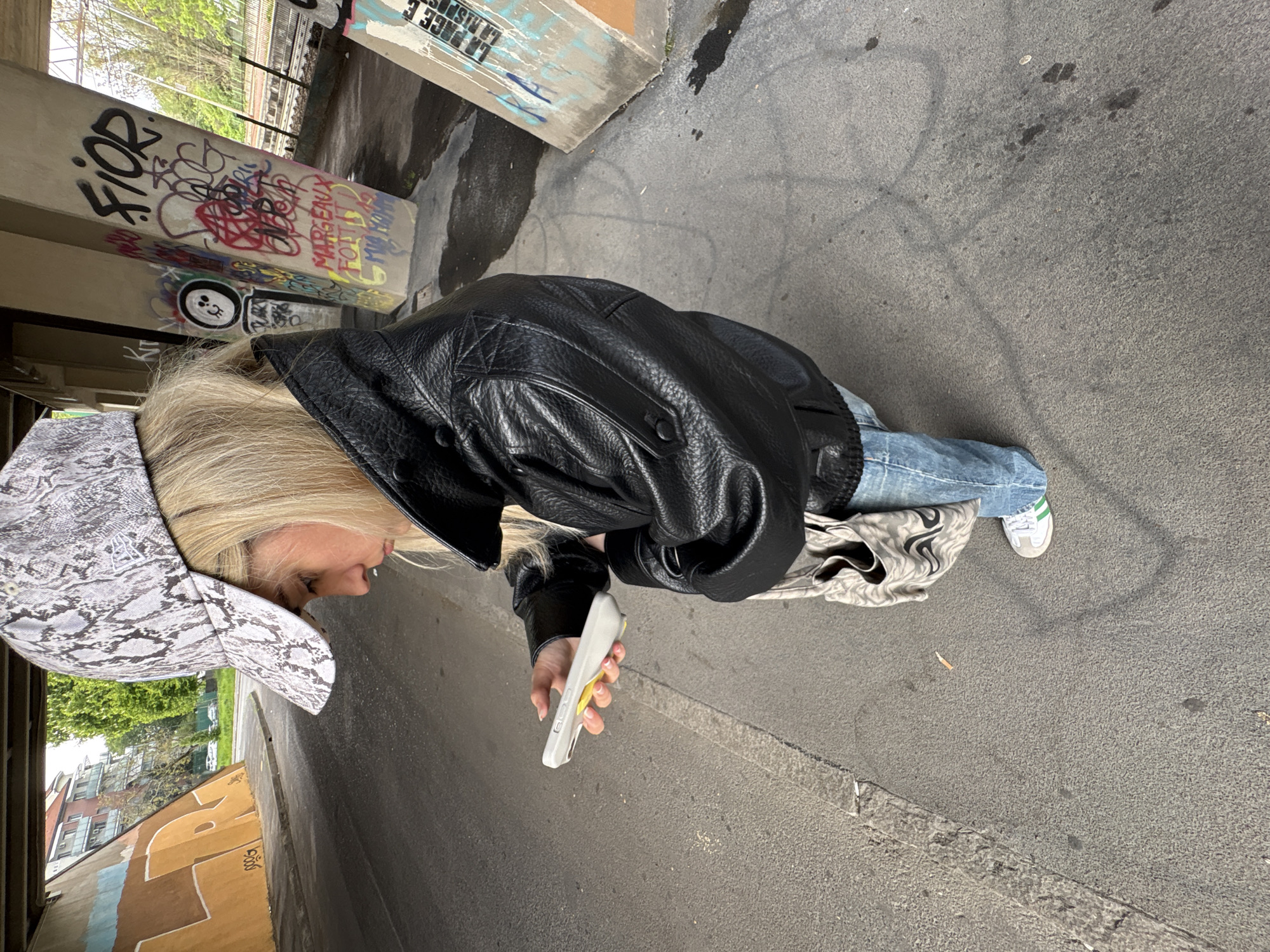 The photograph depicts an individual standing in what appears to be an urban underpass in Milan, Italy. The location is discernible by the presence of graffiti-covered concrete pillars, consistent with the architectural style found in such infrastructure elements. The individual, wearing a black leather jacket, light-wash denim jeans, and white sneakers with green stripes, is holding a white and yellow smartphone. A patterned tote bag rests on the ground next to their feet. The environment is characterized by an asphalt ground surface, suggesting a pedestrian or vehicle access point under a bridge or overpass. The presence of buildings and trees visible in the background indicates the location is within the city limits of Milan.