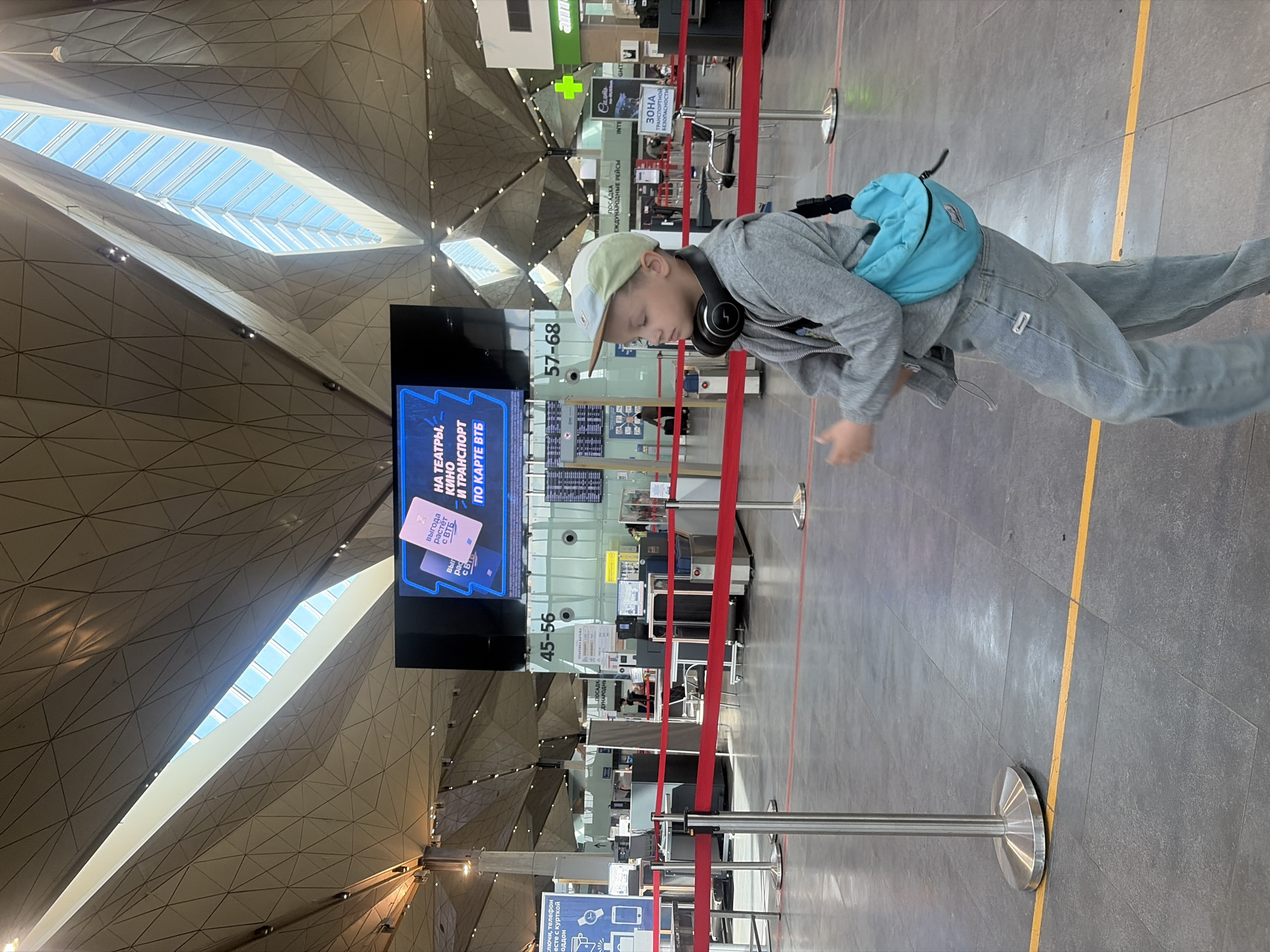 A young person stands in a queue area within the modern terminal of Pulkovo Airport in Saint Petersburg, Russia, during the daytime. The image is rotated 90 degrees counter-clockwise. The individual, appearing to be a boy, wears a light-colored baseball cap, a gray hooded sweatshirt, light blue jeans, and has black over-ear headphones resting around their neck. A bright blue fanny pack is worn across their chest. Their gaze is directed downwards, suggesting a moment of quiet contemplation or waiting.
The background reveals the airport's distinctive geometric ceiling design with prominent skylights that allow natural light to illuminate the space. Red stanchions and barrier ropes delineate the queuing area. Behind these, airport check-in counters or security lanes are visible. A large digital display board shows an advertisement in Russian: "НА ТЕАТРЫ, КИНО И ТРАНСПОРТ ПО КАРТЕ ВТБ" (FOR THEATERS, CINEMA AND TRANSPORT WITH VTB CARD) and "ВЫГОДА РАСТЁТ" (BENEFIT GROWS). Other signs in Russian include "ПОСАДКА МЕЖДУНАРОДНЫЕ РЕЙСЫ" (BOARDING INTERNATIONAL FLIGHTS) next to numbers "45-56" and "57-68," and "ЗОНА ТРАНСПОРТНОЙ БЕЗОПАСНОСТИ" (TRANSPORT SECURITY ZONE). Fragments of text possibly related to security instructions, like "ключи, телефон вместе с курткой и поддоном," are also discernable. The floor is covered with dark gray tiles, featuring a yellow demarcation line.