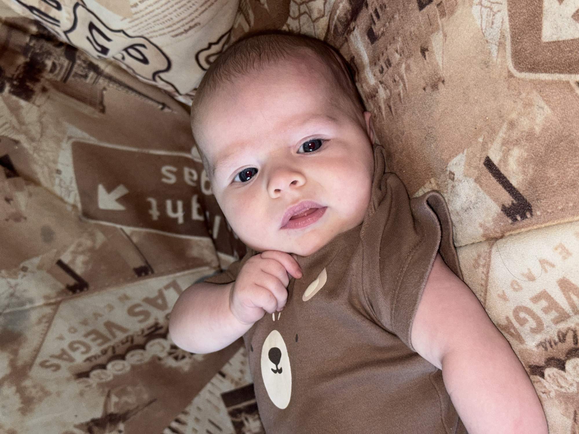 A baby lies on a brown patterned surface. The baby is fair-skinned with dark eyes and is wearing a brown shirt with a bear design. The patterned surface includes text and images, possibly depicting a city such as Las Vegas, Nevada. The location and the city of Chaikovsky, Russia, are not evident in this image.