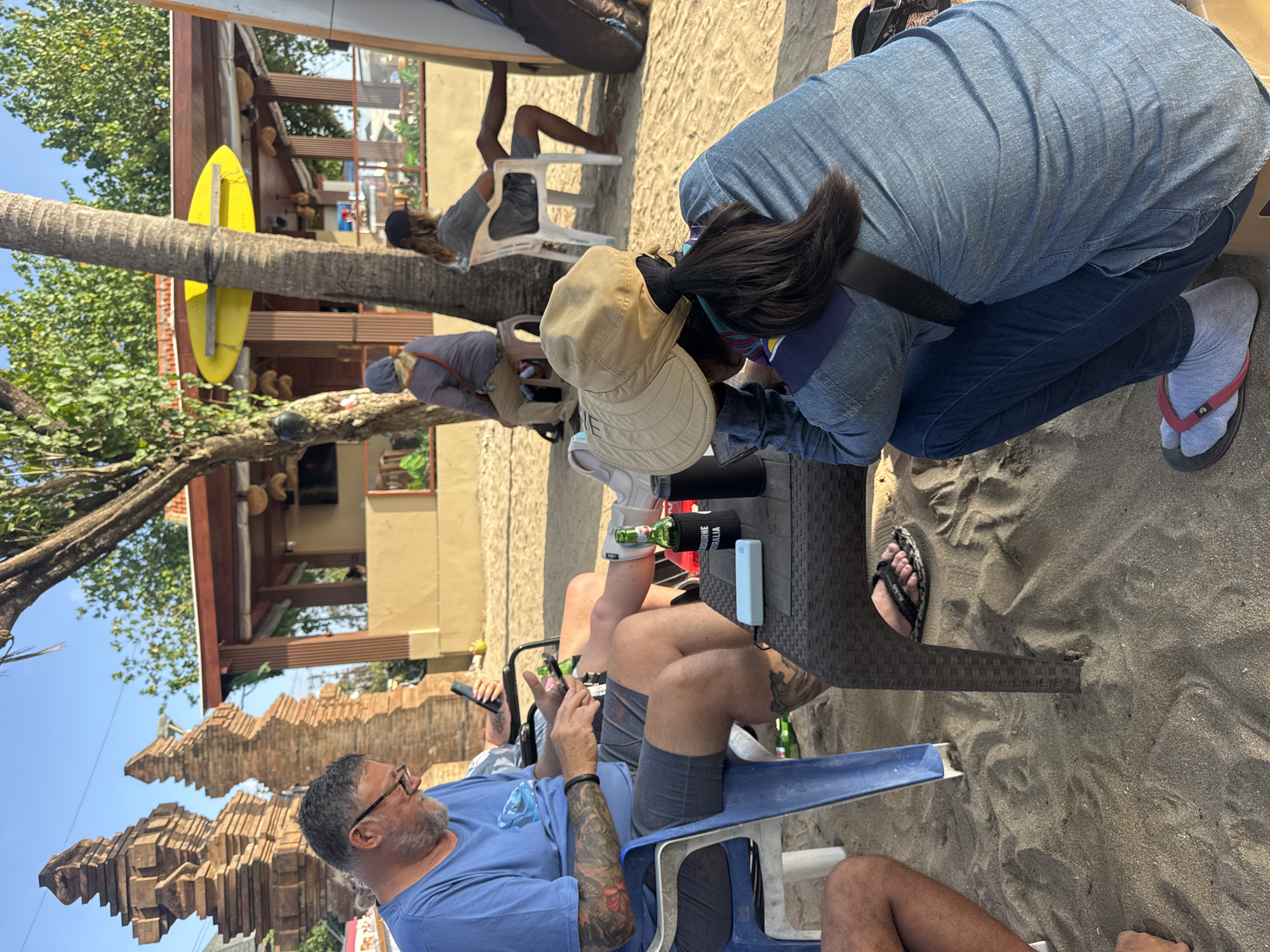 The image depicts a beachside scene in Kuta, Indonesia, where several individuals are seated and engaged with their mobile devices. In the foreground, a person wearing a denim jacket and jeans with a hat is focused on their phone. To the left, a man in a blue t-shirt and shorts is also using his phone. Further back, two other individuals are visible; one is seated on a stool wearing a headscarf, and another is sitting in a white chair. The environment consists of sandy ground, palm trees, and a building with a yellow facade and a red-tiled roof, suggesting it is likely a beach bar or cafe. Surfboards are also visible, indicating a surf-centric location. A beer bottle in a koozie and a power bank are present on a table in the middle ground. The overall atmosphere is relaxed and casual, with people enjoying the outdoors and their technology.