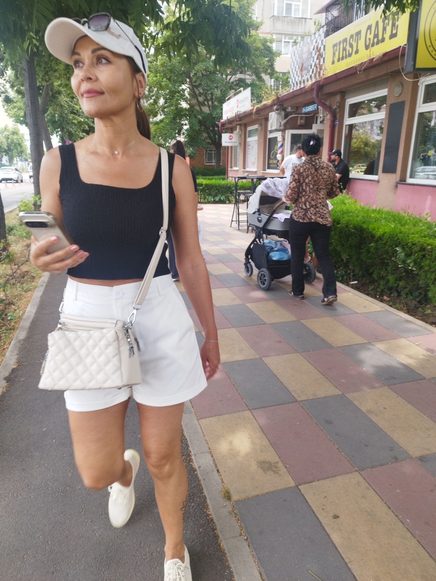 An adult woman wearing a white baseball cap, sunglasses perched on top, a black sleeveless crop top, and white shorts is walking on a sidewalk adjacent to a street. She holds a mobile phone in her left hand and carries a light-colored quilted crossbody bag. The sidewalk is paved with large, rectangular tiles in alternating shades of beige, maroon, and gray. To her right, a row of green bushes borders the sidewalk. Behind the bushes, a building with a pink facade is visible, featuring windows and signage for "FIRST CAFE" and "BARBER". Further in the background, other buildings and trees are present. A person is seen pushing a baby stroller on the sidewalk ahead of the woman, and another person is standing near the building entrance. The scene appears to be set in an urban environment in Constanța, Romania, likely a commercial street.