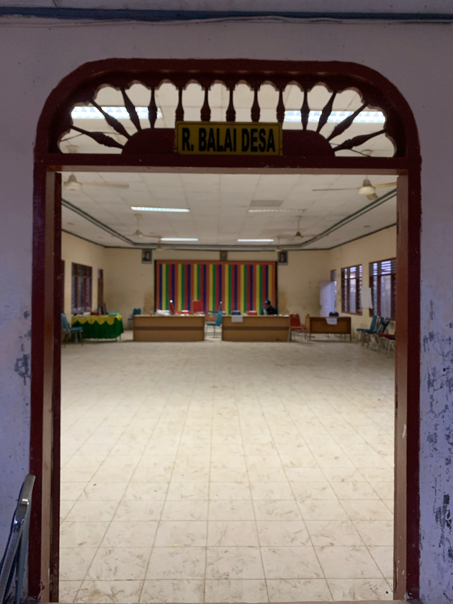 The image shows the entrance to a large room, possibly a community hall or meeting room, within a building in Pantegurah, Indonesia. The entrance is framed by a dark wooden archway with decorative cutouts. Above the entrance, a yellow sign reads "R. BALAI DESA," which translates to "Village Hall."
The room itself is spacious and appears to be tiled with light-colored squares. Fluorescent lights are visible on the ceiling, along with ceiling fans. In the background, there are tables set up, some with chairs. A colorful striped backdrop, featuring vertical bands of red, yellow, green, blue, and purple, is visible behind one of the tables. On one of the tables, there is a laptop and a person sitting. Several chairs are arranged around the tables and in other areas of the room. Windows with slatted coverings are present along the walls. The overall impression is that of a functional, multi-purpose indoor space.