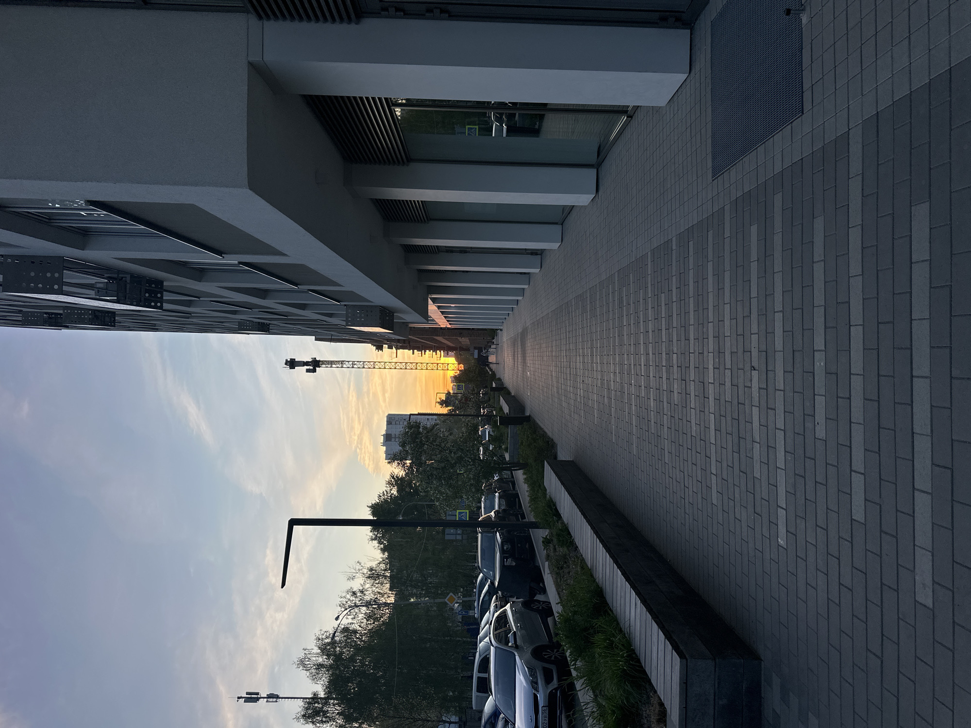 The image shows a street scene in Yekaterinburg, Russia, taken at ground level. The camera points down a paved walkway lined with modern, white buildings on the left. The buildings have a repeating pattern of rectangular windows and architectural supports. To the right of the walkway is a raised curb separating it from a row of parked cars. Beyond the cars are trees and bushes. In the distance, a crane is visible against the backdrop of a sunset. The sky is a mix of blue and orange hues. Street lights are also visible. The light suggests it is either sunrise or sunset.