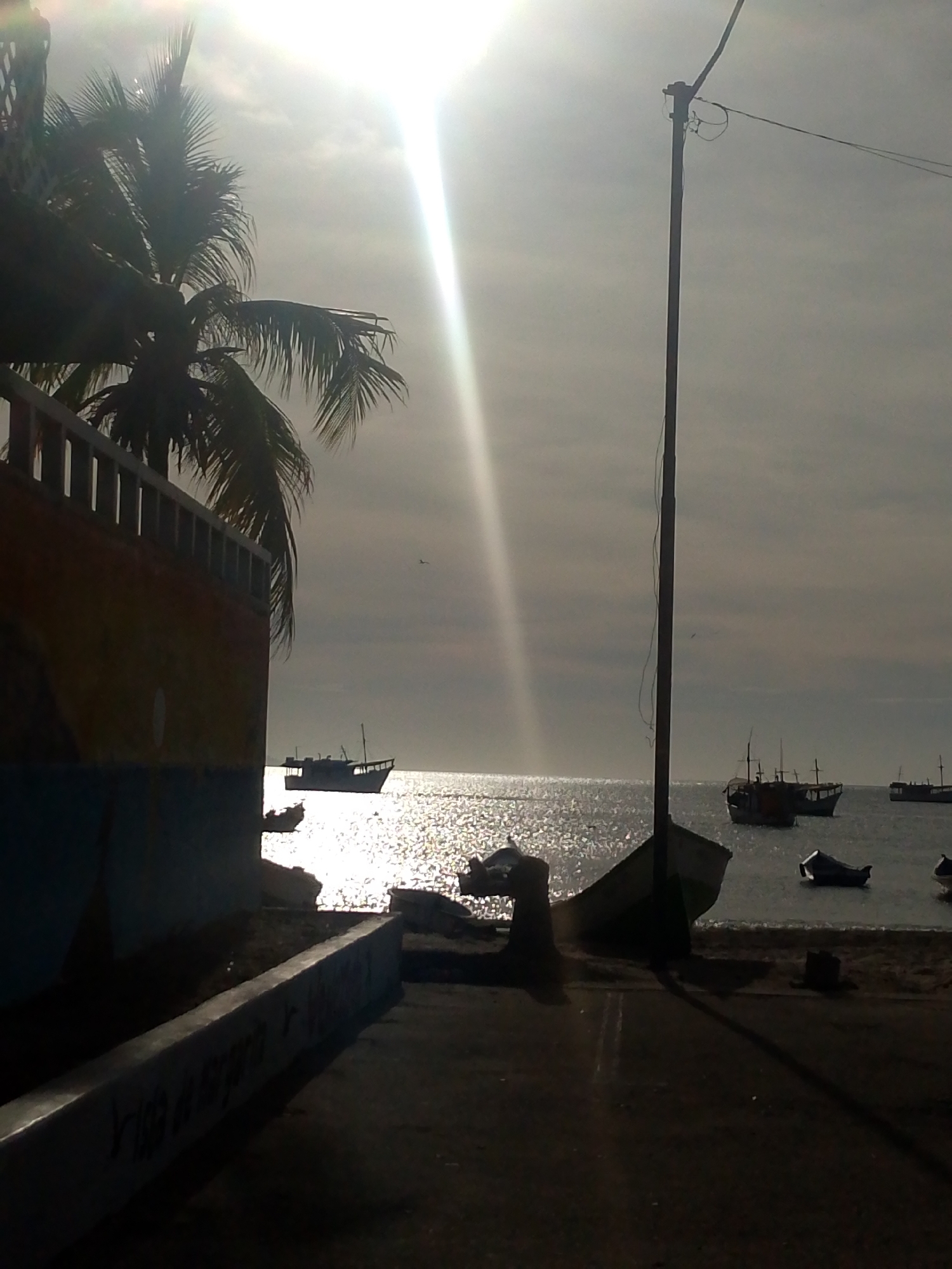 The image depicts a coastal scene in Juan Griego, Venezuela. The foreground is dominated by a dark, paved walkway with a low concrete barrier on the left, and a palm tree and partially visible building also on the left. A utility pole stands on the right edge of the walkway. The background features a shimmering ocean with several small boats anchored in the water. The sky is overcast, with the sun appearing as a bright glare. The overall impression is one of a quiet, possibly early morning or late afternoon scene at a harbor.