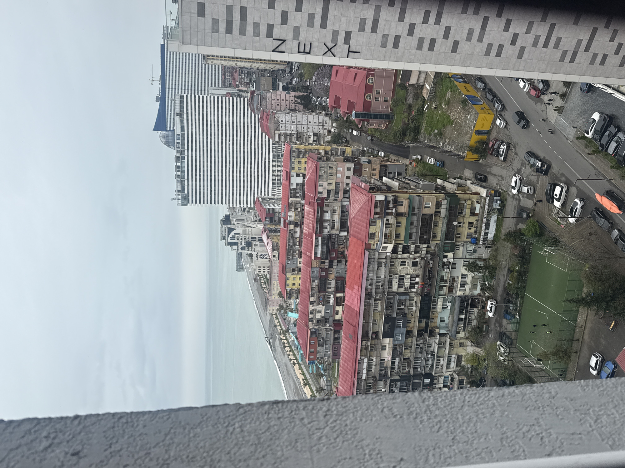The image shows an aerial view of a cityscape, likely Batumi, Georgia, with the Black Sea visible on the left. The buildings are densely packed, with a mix of architectural styles, including taller modern structures and older buildings with red roofs. A street runs parallel to the coast, with cars parked along the sides and traffic moving. There is a green space that appears to be a soccer field with people on it. The sky is overcast. The upper portion of the image contains lettering that reads "NEXT."