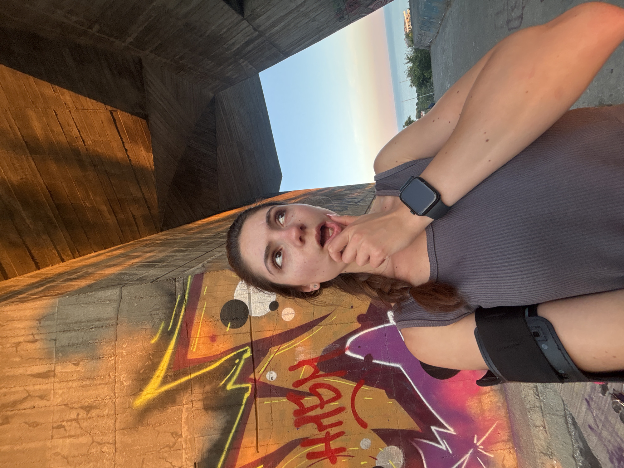 A young woman with brown hair in a braid is standing outdoors, partially shaded by a concrete structure with graffiti on the wall. She is wearing a grey tank top and a black sports watch on her left wrist. Her right hand is raised to her mouth, and she appears to be looking up and to the side with a thoughtful expression. The background shows a clear blue sky fading into a pink and orange sunset over the sea, with some greenery and buildings visible in the distance. The location is Varna, Bulgaria, likely near a coastal area or a bridge, given the concrete structure and the sea view.