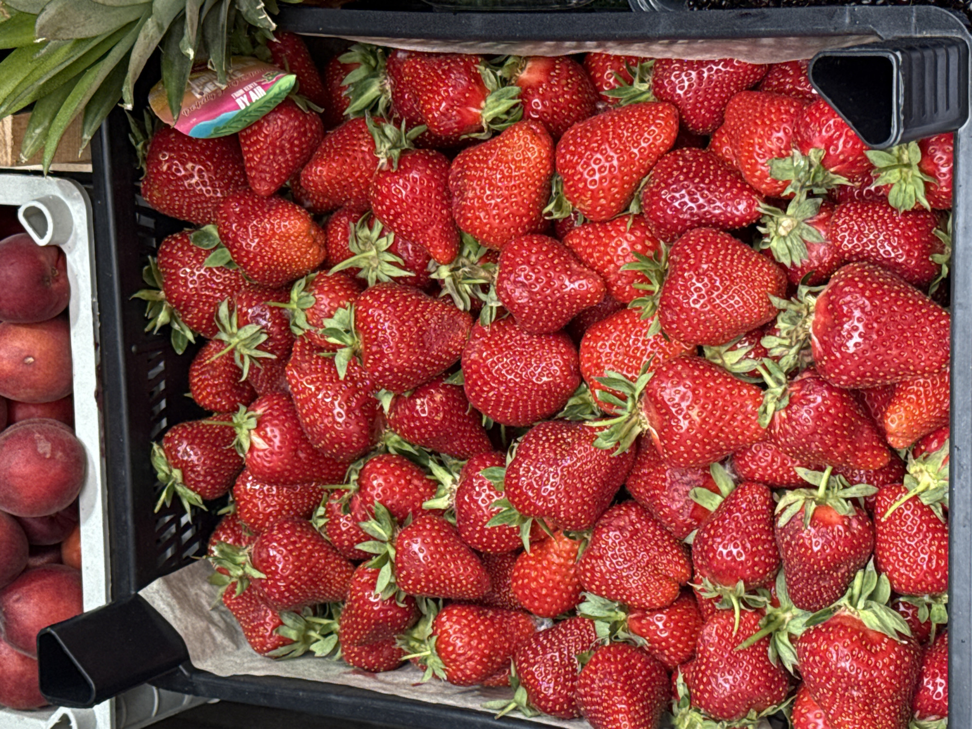 The image shows a close-up view of a black plastic crate filled with ripe strawberries. The strawberries are a deep red color, with visible seeds on their surface and green stems. Some of the strawberries are larger than others, and they are packed tightly together. In the background, partially visible, are some peaches and a pineapple. The overall scene suggests a market or a produce stall. No specific location in Saint Petersburg, Russia is discernible from the image alone.