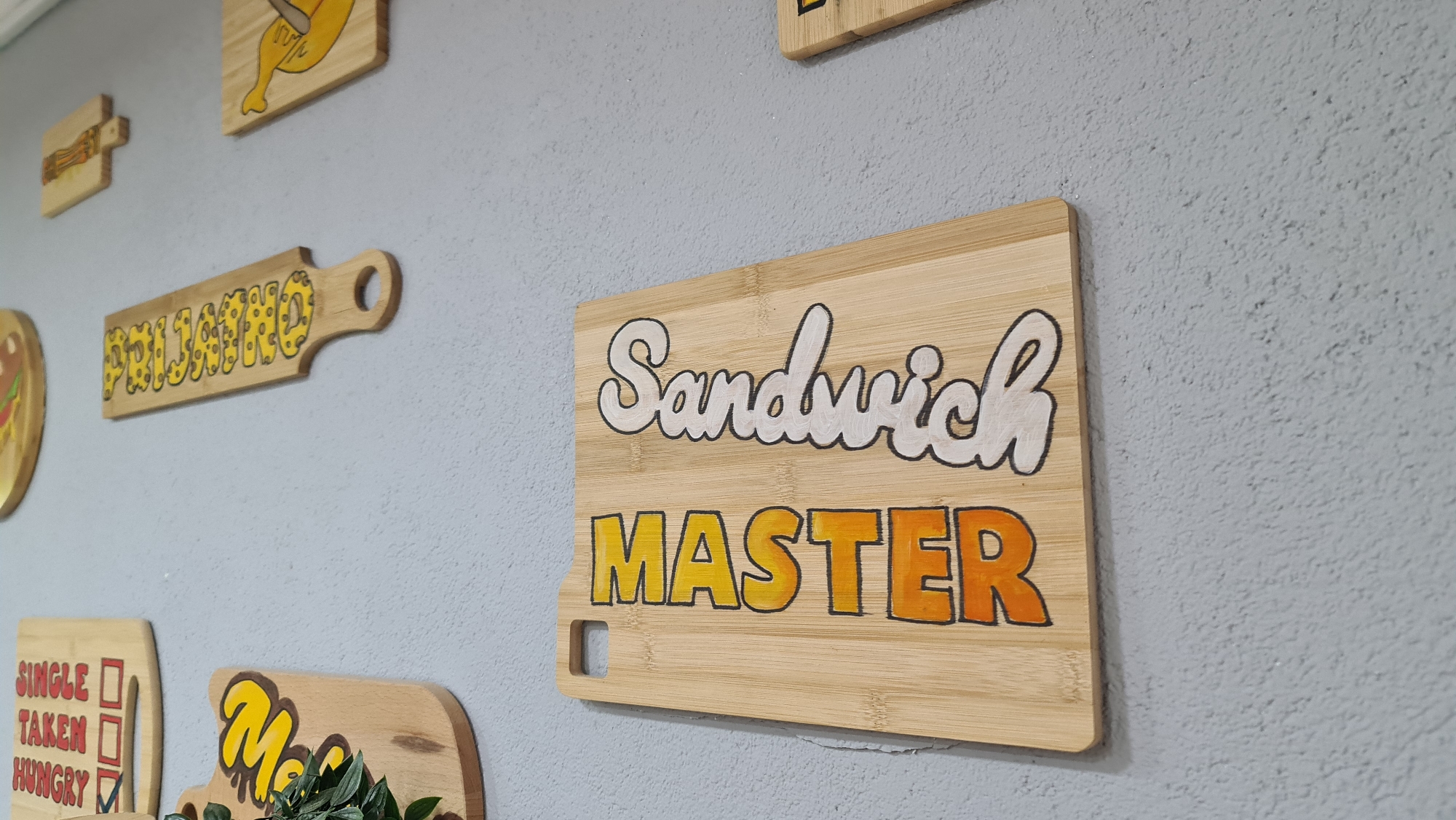 The image depicts a wall decoration consisting of several wooden plaques. One plaque, prominently displayed on the right, is rectangular with rounded corners and made of bamboo. It reads "Sandwich MASTER" in stylized lettering, with "Sandwich" written in white and "MASTER" in a gradient from yellow to orange. This plaque has a small square hole in the lower left corner.
Other plaques can be seen on the wall, including one that says "PRIJATNO" in yellow lettering on a long, narrow plaque with a handle. Another shows a checklist with "SINGLE," "TAKEN," and "HUNGRY," with the "HUNGRY" box checked. Additional plaques are present with partial visibility.
The background is a textured gray wall.
There is no information in the image about a location in Budva, Montenegro.