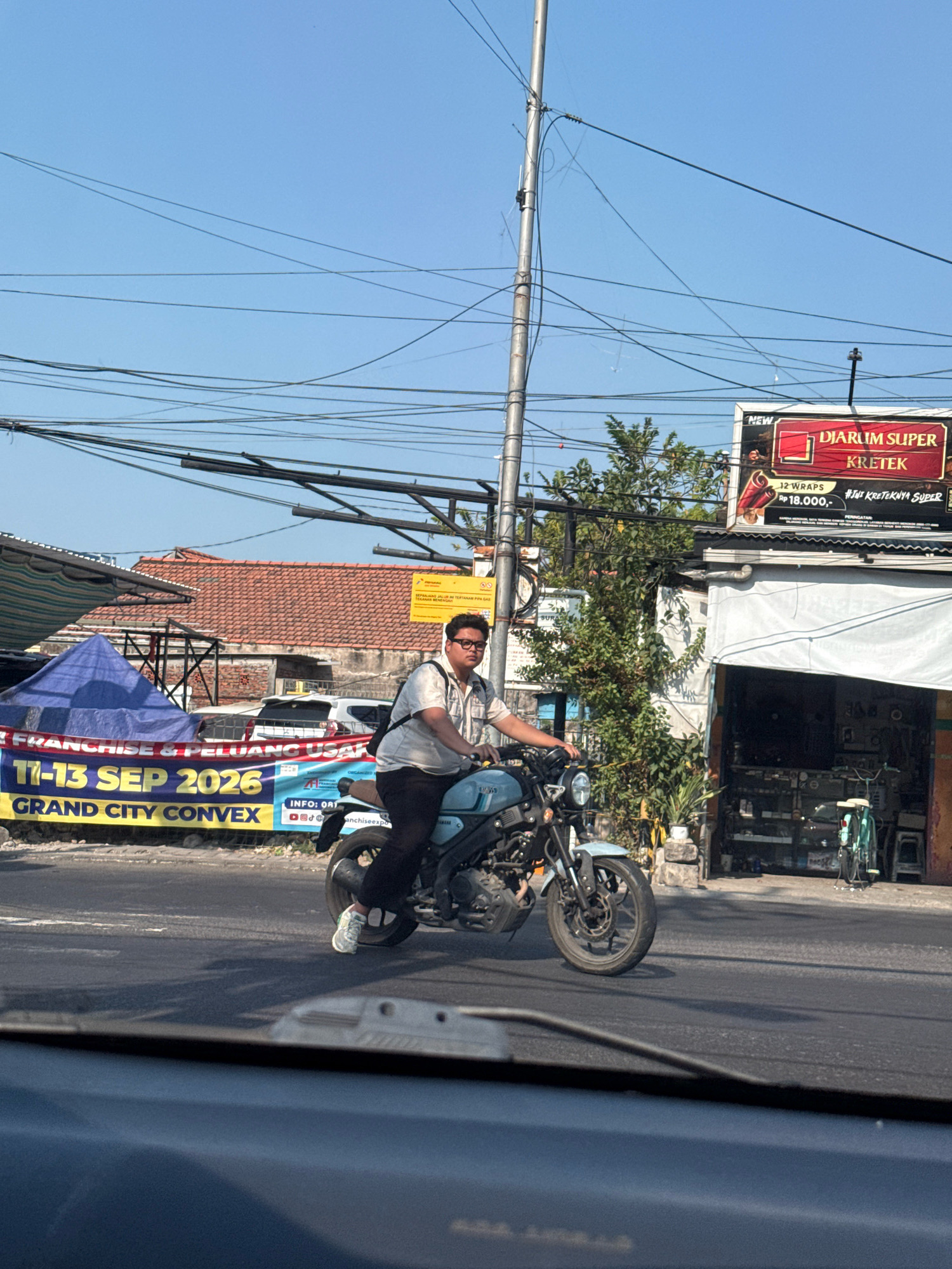 A young man wearing glasses and a light-colored collared shirt is riding a light blue motorcycle on a paved road. He is facing the viewer's right, and his left leg is bent at the knee, while his right leg is extended down toward the ground. The man is wearing black pants and white sneakers. The motorcycle has a black seat and handlebars, a silver front fender, and a silver exhaust pipe. The road is asphalt, and there is a white line painted on it. In the background, there is a building with a red tile roof and a blue tarp. There are also power lines strung across the sky. A banner across the road reads "FRANCHISE & PELUANG USAHA 11-13 SEP 2026 GRAND CITY CONVEX". Another sign above a shop reads "NEW DJARUM SUPER KRETEK". The scene is likely in Surabaya, Indonesia, given the context of the banner and the style of the buildings and signage.