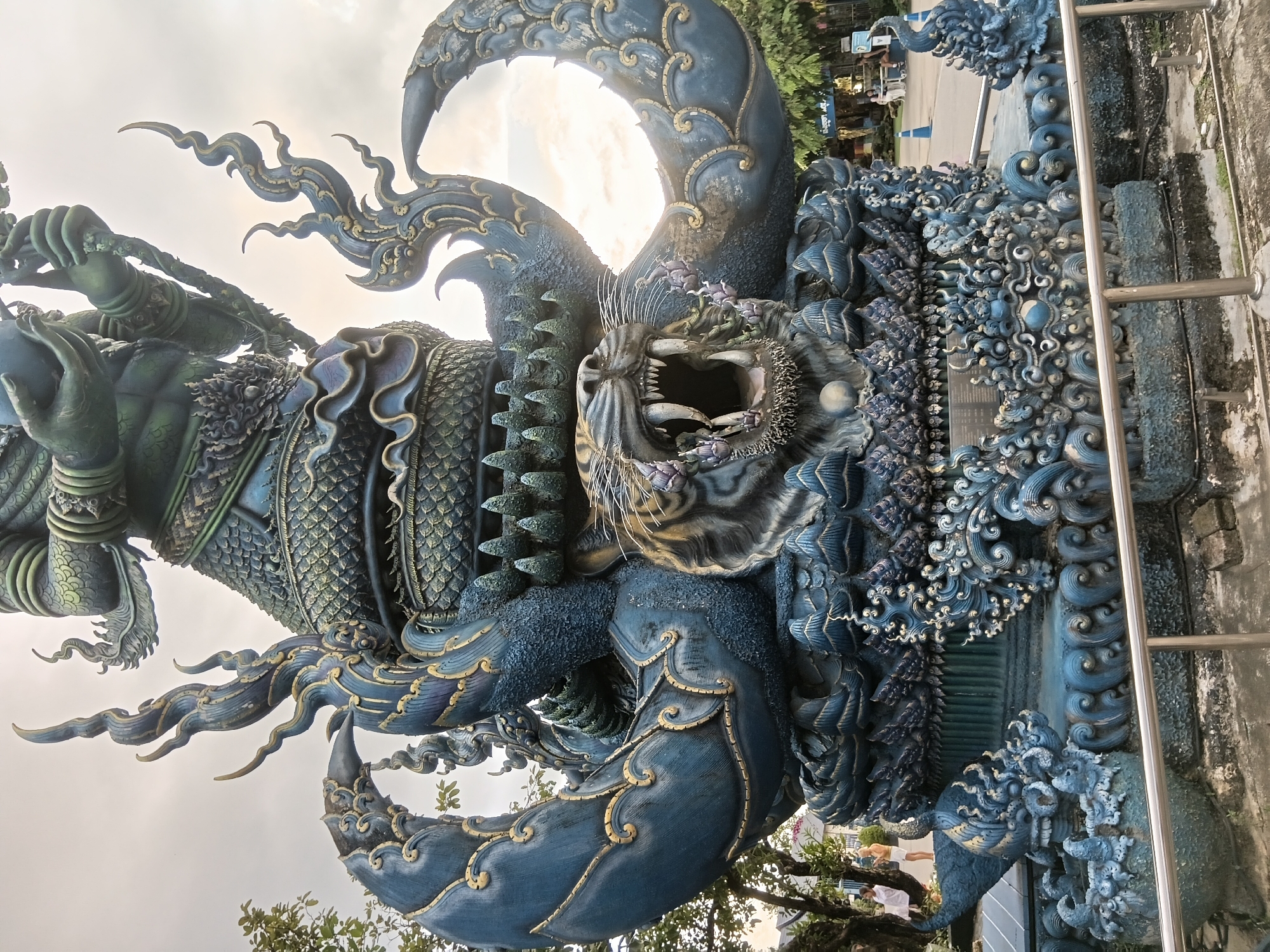 The image shows a large, ornate blue statue with gold accents, depicting a mythical creature that appears to be a combination of a serpent and a tiger. The tiger's head, with its mouth agape and sharp teeth visible, is a prominent feature. The serpent's body coils around the tiger, with scaled textures and decorative elements. The statue is set against a cloudy sky, with some trees and distant buildings visible in the background. The overall impression is one of grandeur and intricate artistry. This statue is located at the Wat Rong Suea Ten (Blue Temple) in Chiang Rai City Municipality, Thailand.