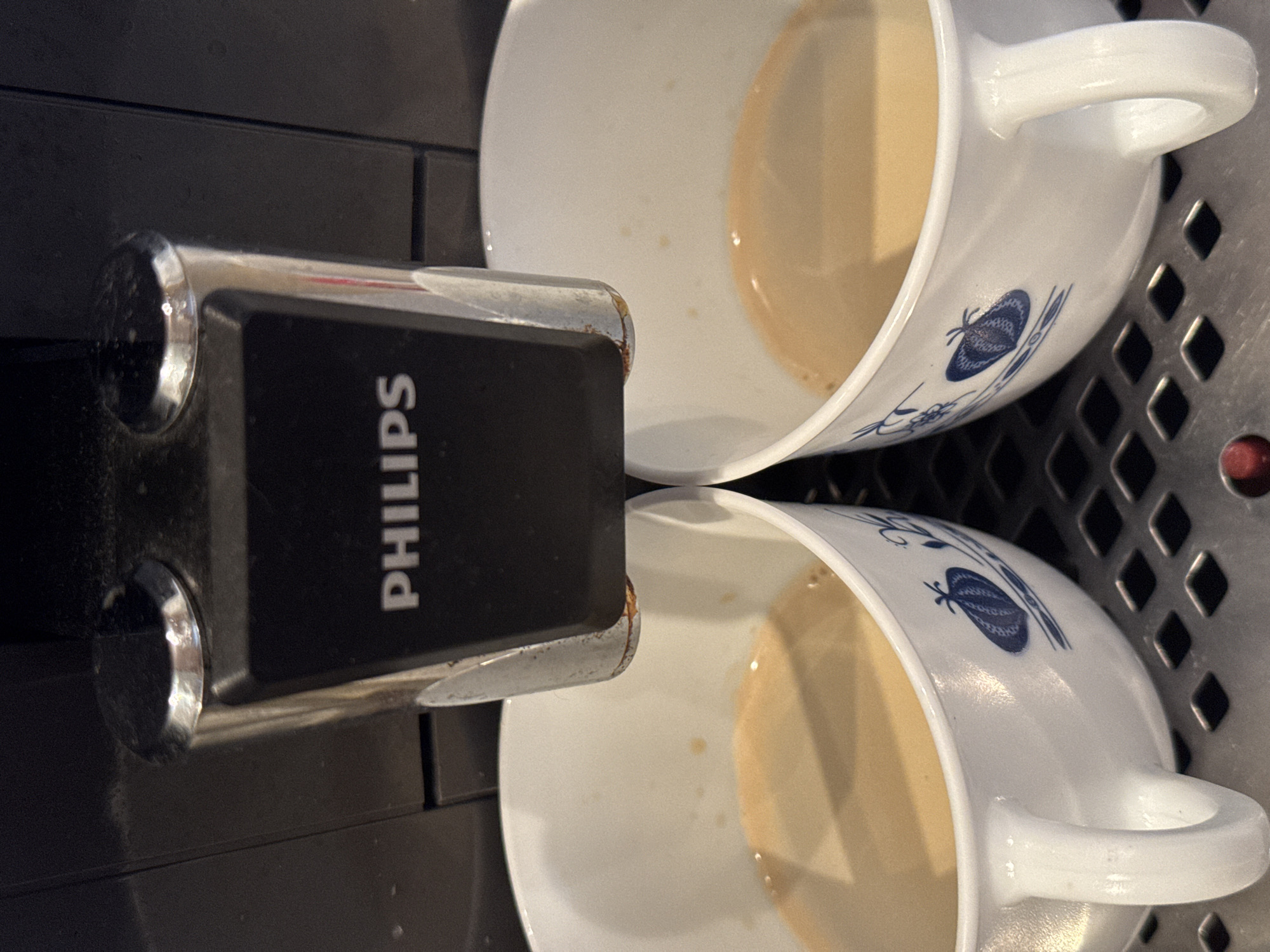 The image, rotated 90 degrees clockwise, presents a close-up view of two white ceramic mugs filled with a light brown beverage, positioned beneath a coffee machine dispenser.
The central subject is a black and silver coffee machine component, likely a double spout, from which the liquid has been or is being dispensed. The word "PHILIPS" is printed vertically in white capital letters on the black surface of this component. The silver sections of the dispenser appear somewhat worn or smudged.
Below the dispenser, two identical white ceramic mugs are visible, each containing a light brown liquid, suggestive of coffee with milk or cream. Both mugs feature a blue decorative pattern, which includes a prominent butterfly motif and other abstract floral or leafy designs. The liquid in the mugs shows small bubbles on its surface.
The mugs are resting on a dark-colored metal grate, which is part of a drip tray. A small, circular red element is partially visible on the far right edge of this grate. The scene suggests an indoor setting, possibly a kitchen or office break area. No people are visible, and the time of day is not discernible.