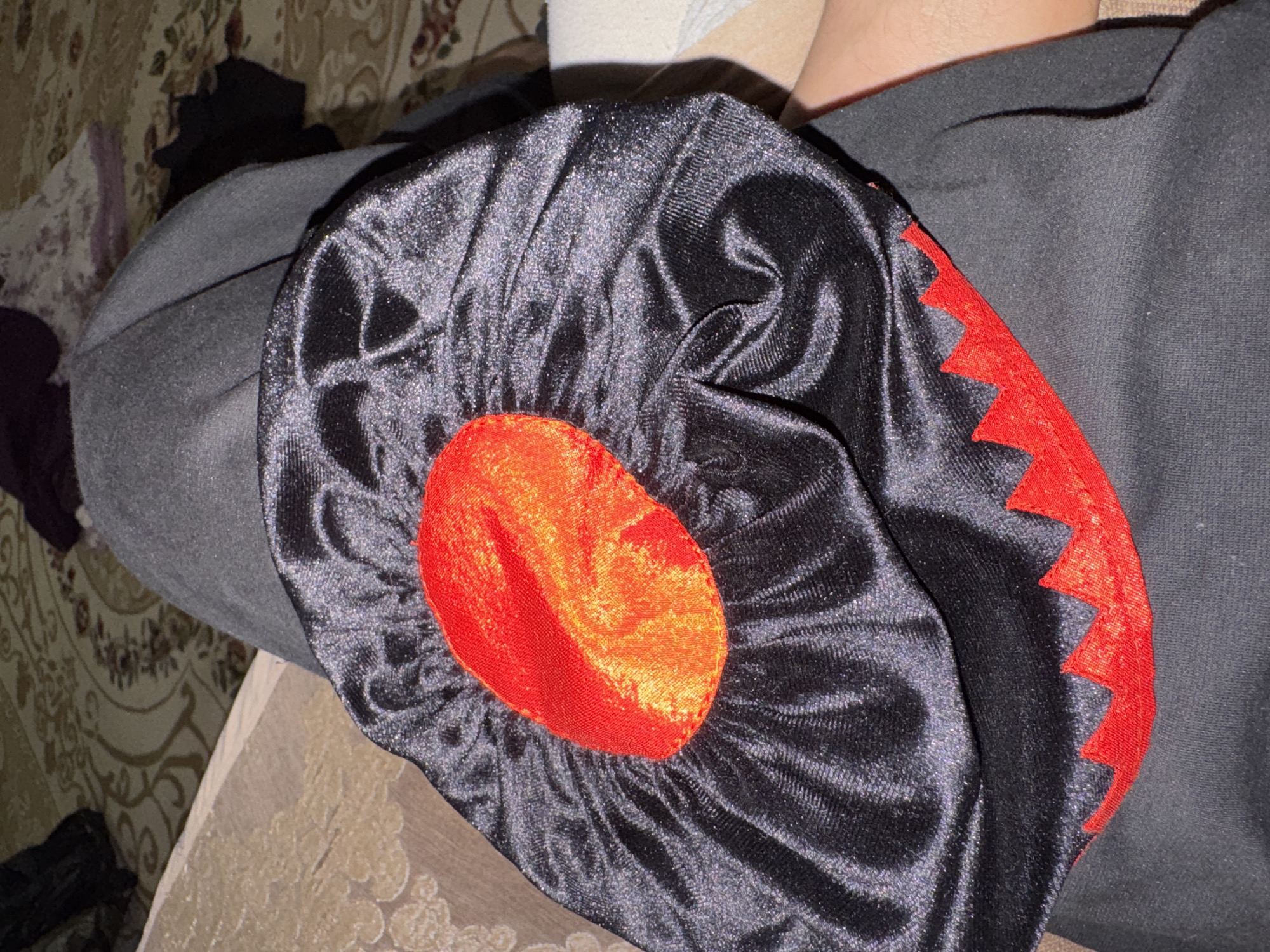 A close-up shot shows a black satin costume piece with a red and orange circular emblem in the center, resting on gray fabric. The costume piece has a jagged red trim along its bottom edge. The background consists of a patterned carpet and a hint of skin. The image is taken in the city of Kurchaloy, Russia.