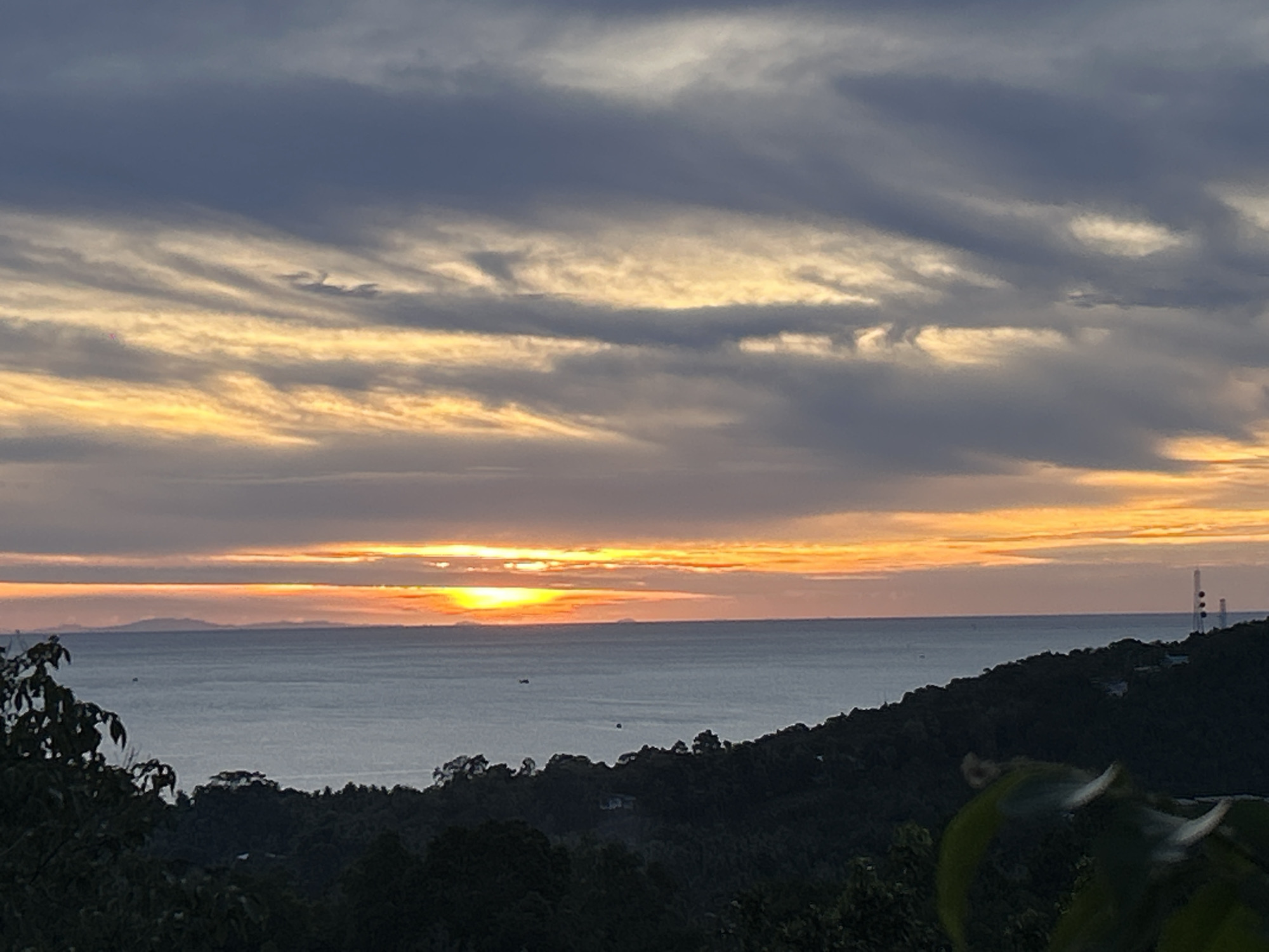 The image depicts a sunset over the sea in Baan Sri Thanu, Thailand. A low sun is visible above the horizon, its light reflected on the water. There are dark islands or landmasses in the distance on the left side. Above the sea, layers of clouds are streaked with gold from the setting sun. The foreground shows a dark, tree-covered slope. A communication tower is visible on a distant hillside. There are a few small boats scattered on the water's surface.
