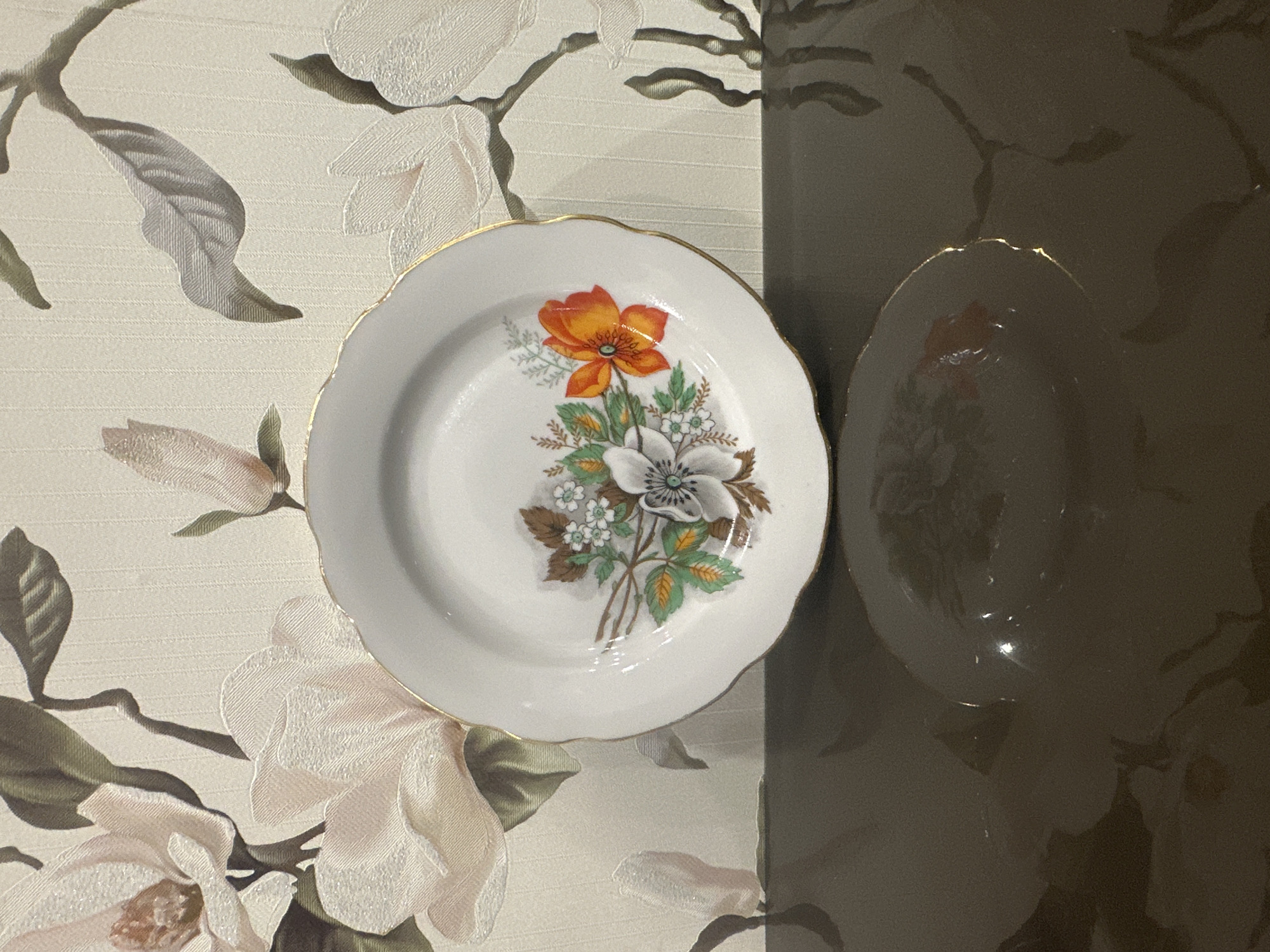 A close-up, eye-level shot features a white ceramic plate with a floral pattern and a gold rim, centered on a dark, reflective surface that appears to be a table. The plate's design includes several flowers, prominently a white one with a dark center and an orange one with visible stamens, surrounded by green leaves and branches. The background is a textured wallpaper with a repeating pattern of large, pale pink magnolias and green leaves. The lighting is soft, casting gentle reflections on the plate and the surface below. The image composition is balanced, with the plate as the clear focal point. This image was not taken in Bryansk, Russia.