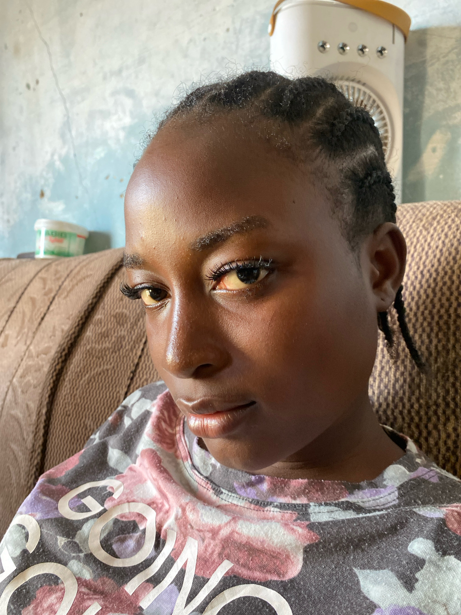 The image shows a close-up, eye-level portrait of a young Black person with dark skin and braided hair. The subject is looking towards the camera, with their head tilted slightly. They are wearing a grey t-shirt with a floral pattern in pink, purple, and white, and large white lettering that appears to read "GOOD VIBES". The background includes a textured brown sofa, a wall with a light blue wash and visible cracks, and a small white and yellow portable fan. A white container with a green label is visible on a surface behind the subject's left shoulder. The lighting suggests an indoor setting. The person's eyes are open, and they appear to have false eyelashes. The overall impression is a casual, everyday scene. The location is identified as Sabon Tasha, Nigeria.