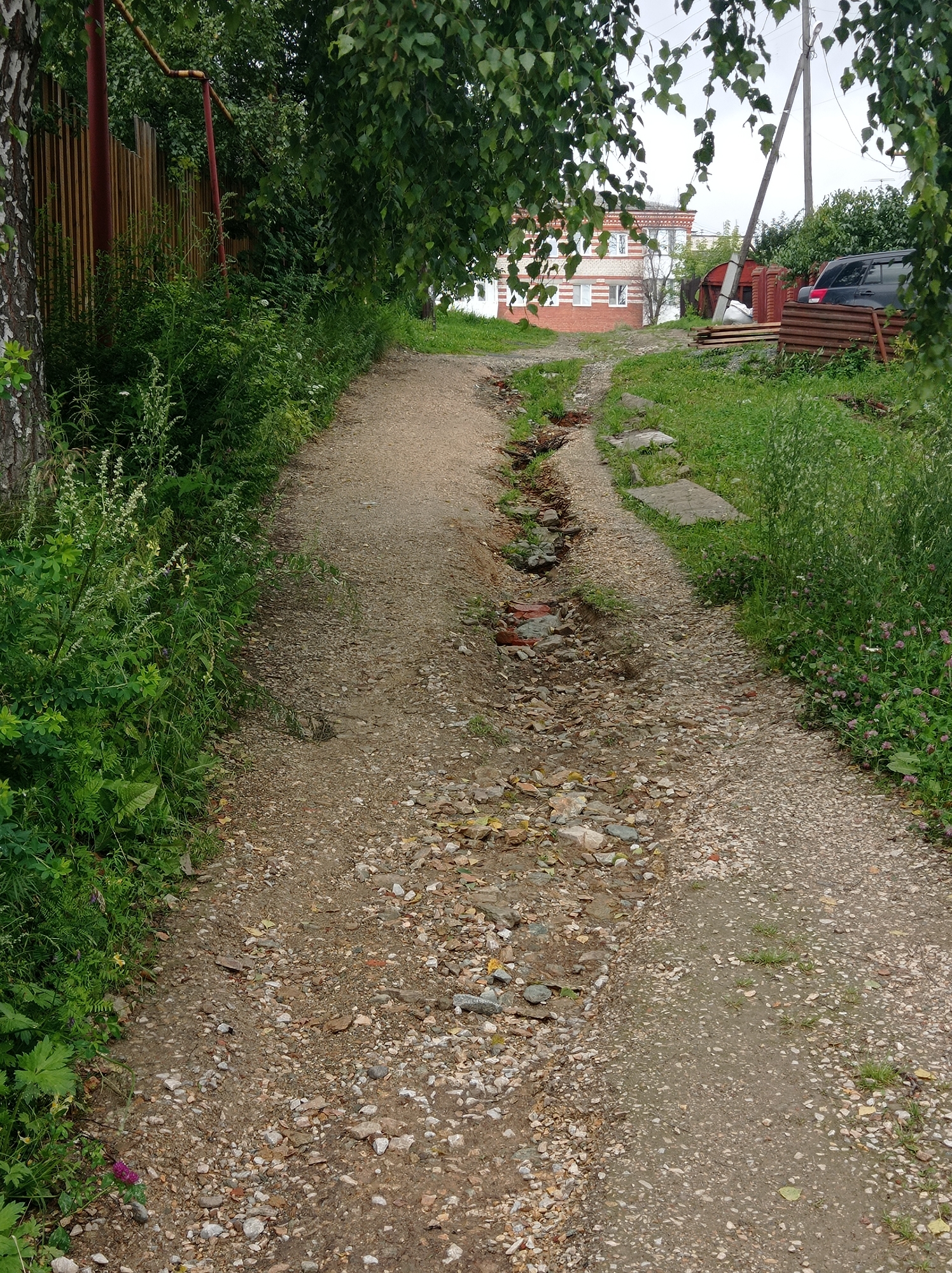 The image depicts a rough, unpaved road winding uphill. The road is a mix of dirt and gravel, with noticeable ruts and erosion in the center, revealing some broken bricks and rocks. Lush green vegetation, including tall grass and bushes, borders the left side of the road, with a birch tree trunk visible on the far left. A wooden fence, painted a reddish-brown, runs along the left side, partially obscured by foliage. On the right side, the vegetation is also dense, with some small purple flowers visible. Further up the road, a two-story building with red brick and white window frames is visible, suggesting it is a residential structure. A dark-colored SUV is parked to the right of the building, next to a pile of wooden planks and a red, corrugated metal structure. A utility pole stands near the building. The overall scene suggests a rural or semi-rural setting within the city of Degtyarsk, Russia, characterized by unmaintained infrastructure and abundant plant life.