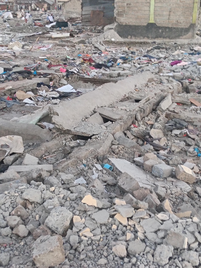 Extensive rubble field dominates the scene, composed of concrete chunks, small stones, and debris. Remnants of structures suggest prior buildings, with bits of walls and foundations visible. Scattered clothing and personal items lie amidst the wreckage. Two blurred figures can be seen at the top left, possibly inhabitants of the place. One building, partially intact, stands at the top right. The overall impression is one of destruction and displacement.