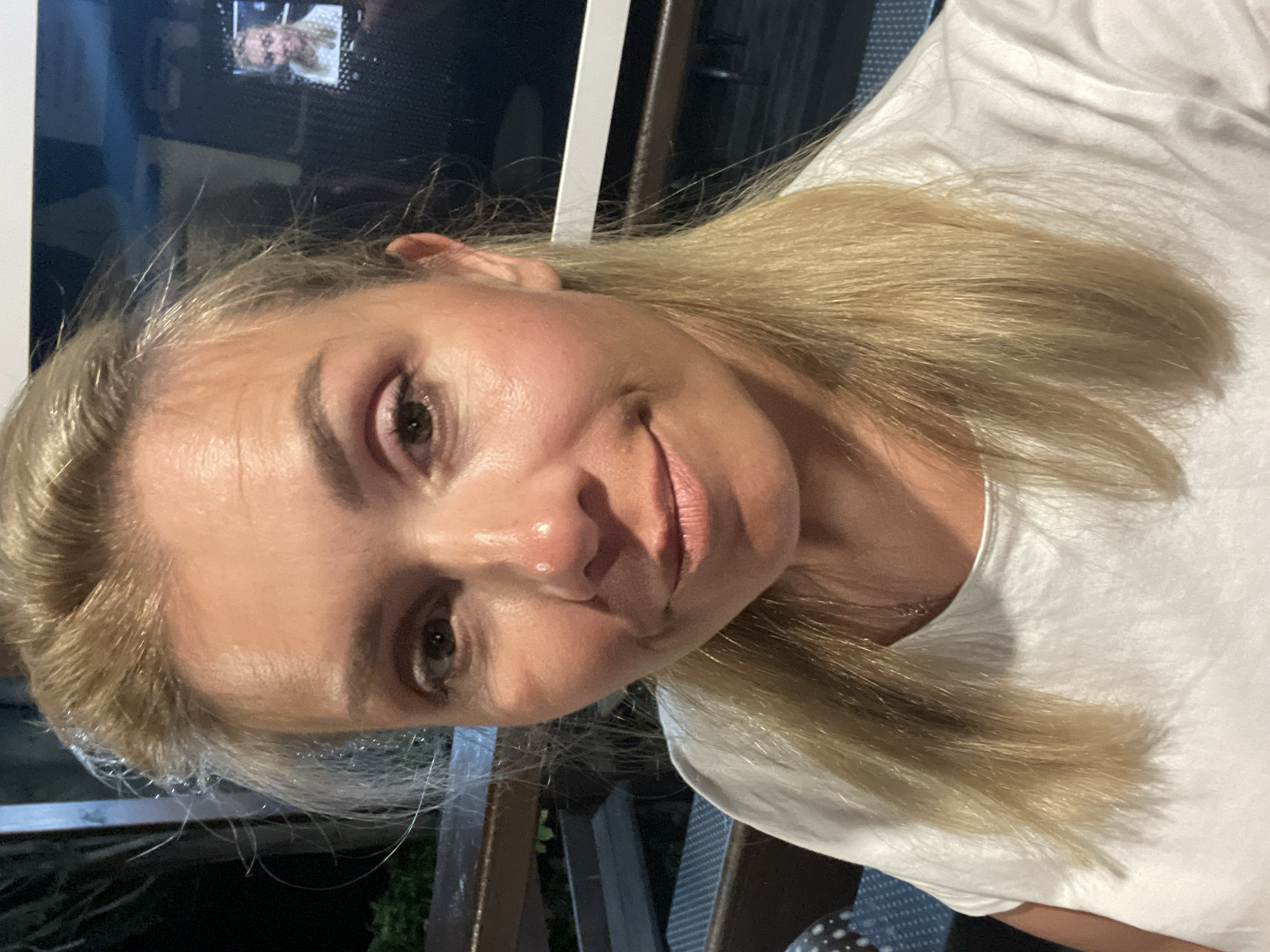 A woman with blonde hair, wearing a white t-shirt, takes a selfie. She is looking towards the camera with a slight smile. In the background, there is a screen displaying a smaller image of the same woman. The environment appears to be indoors, possibly a lounge area, with furniture and dim lighting. The overall scene suggests a casual setting.