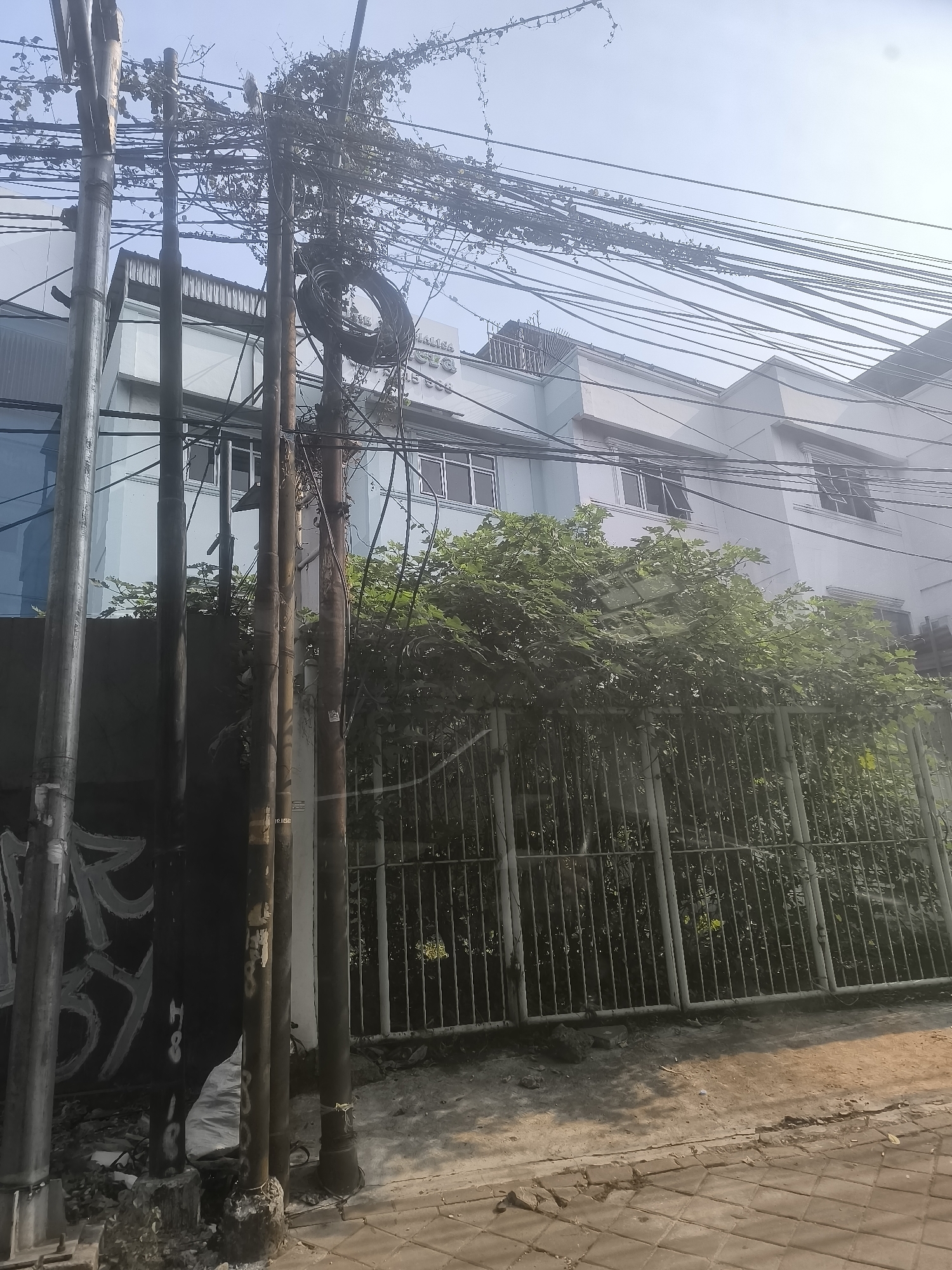 The image depicts a street scene in Tangerang, Indonesia, characterized by a dense network of overhead electrical wires, many of which are tangled with vegetation. In the foreground, several utility poles stand prominently, some appearing weathered and covered in graffiti. Behind the poles and wires, a light-colored building with multiple windows is visible. A metal fence runs along the property line, separating the street from the building's grounds, which are overgrown with trees and bushes. The ground in the foreground consists of a paved walkway and a dirt area littered with debris. The sky is overcast.