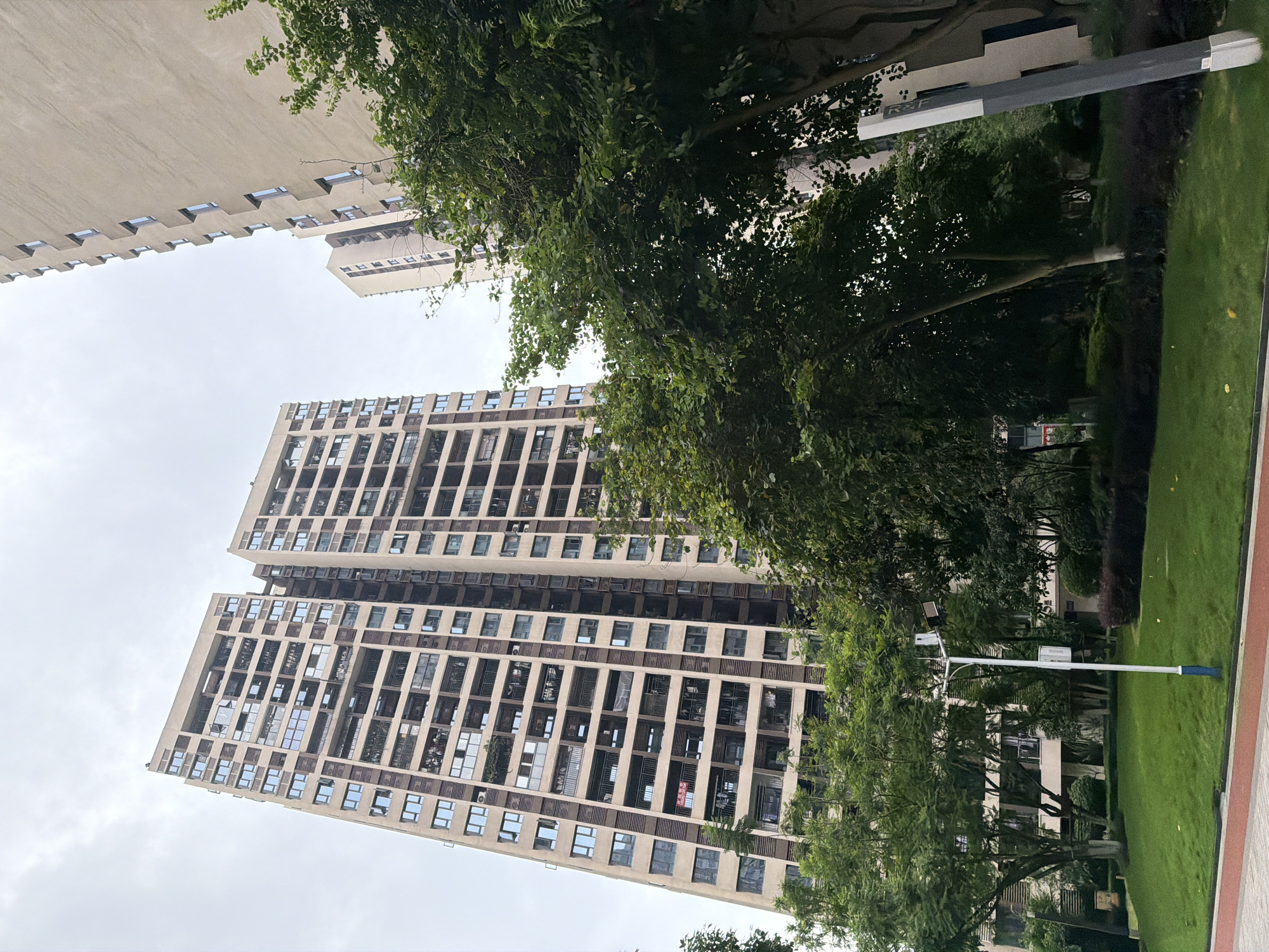 The image captures a view of high-rise apartment buildings in the Chongqing High-tech Zone, China. The buildings are modern, with numerous balconies and windows. Lush green trees and manicured lawns surround the buildings, suggesting a well-maintained residential area. A lamppost stands in the foreground, illuminating the grassy area. The sky is overcast, indicating a cloudy day. There are no people visible in the image, and the scene is tranquil. The architecture suggests a densely populated urban environment, while the greenery offers a touch of nature within the city.