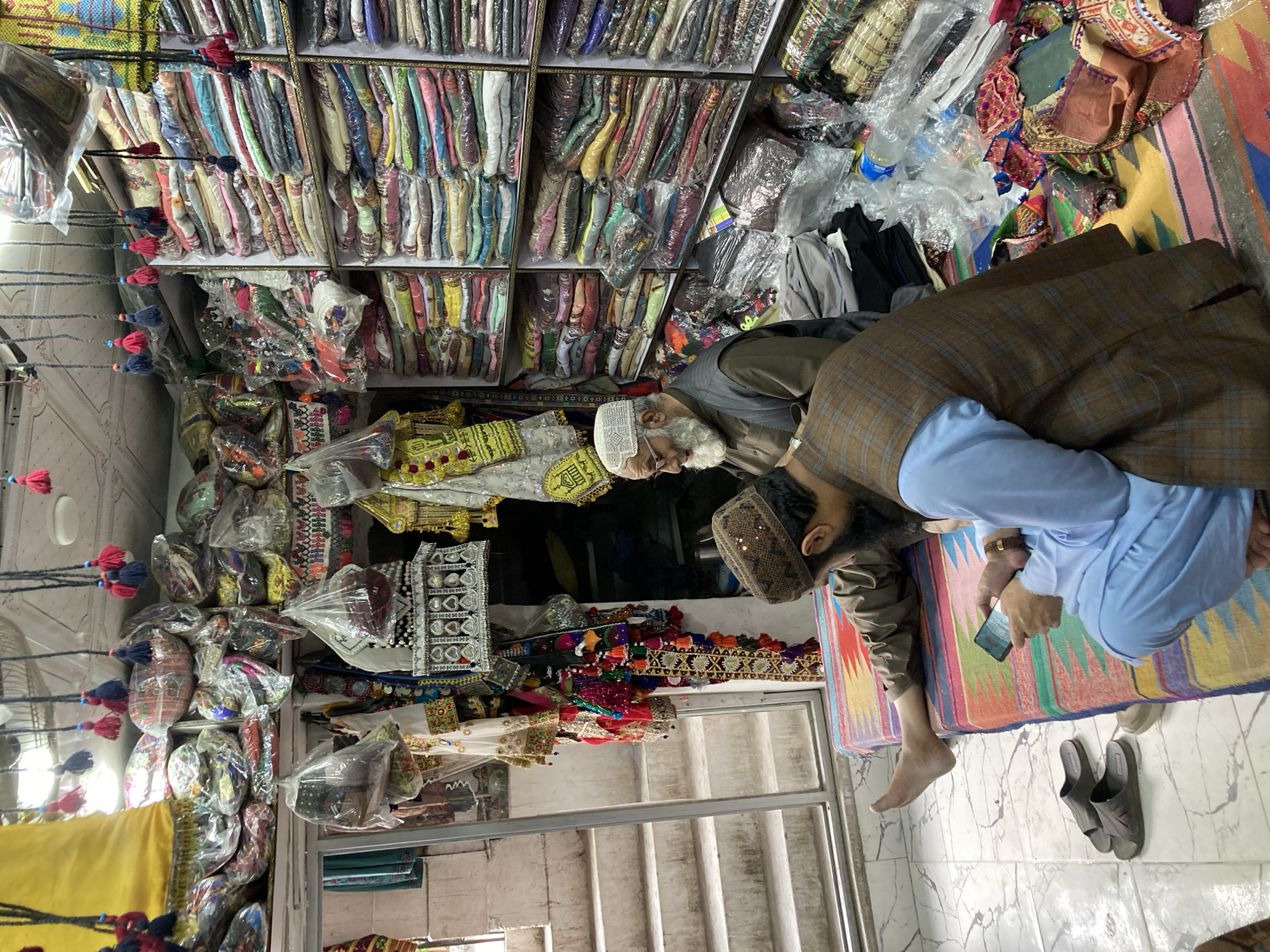 An indoor market in Quetta City Tehsil, Pakistan, shows shelves stocked with textiles and embellished items. Two men are present: one wearing a white skullcap and eyeglasses, and the other in a brown plaid jacket and blue shalwar kameez. The latter is holding a mobile phone. A colorful patterned rug lies on the floor, near a pair of sandals. Stairs are visible in the background.